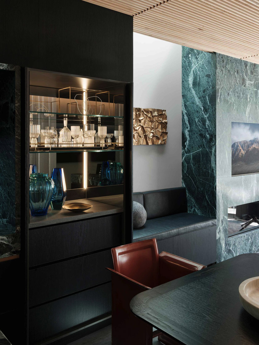 A stylish dining area in The Glasshouse by Nina Maya Interiors, showcasing a black marble table, leather chairs, and a glass display cabinet with decorative glassware, highlighted by green marble accents.