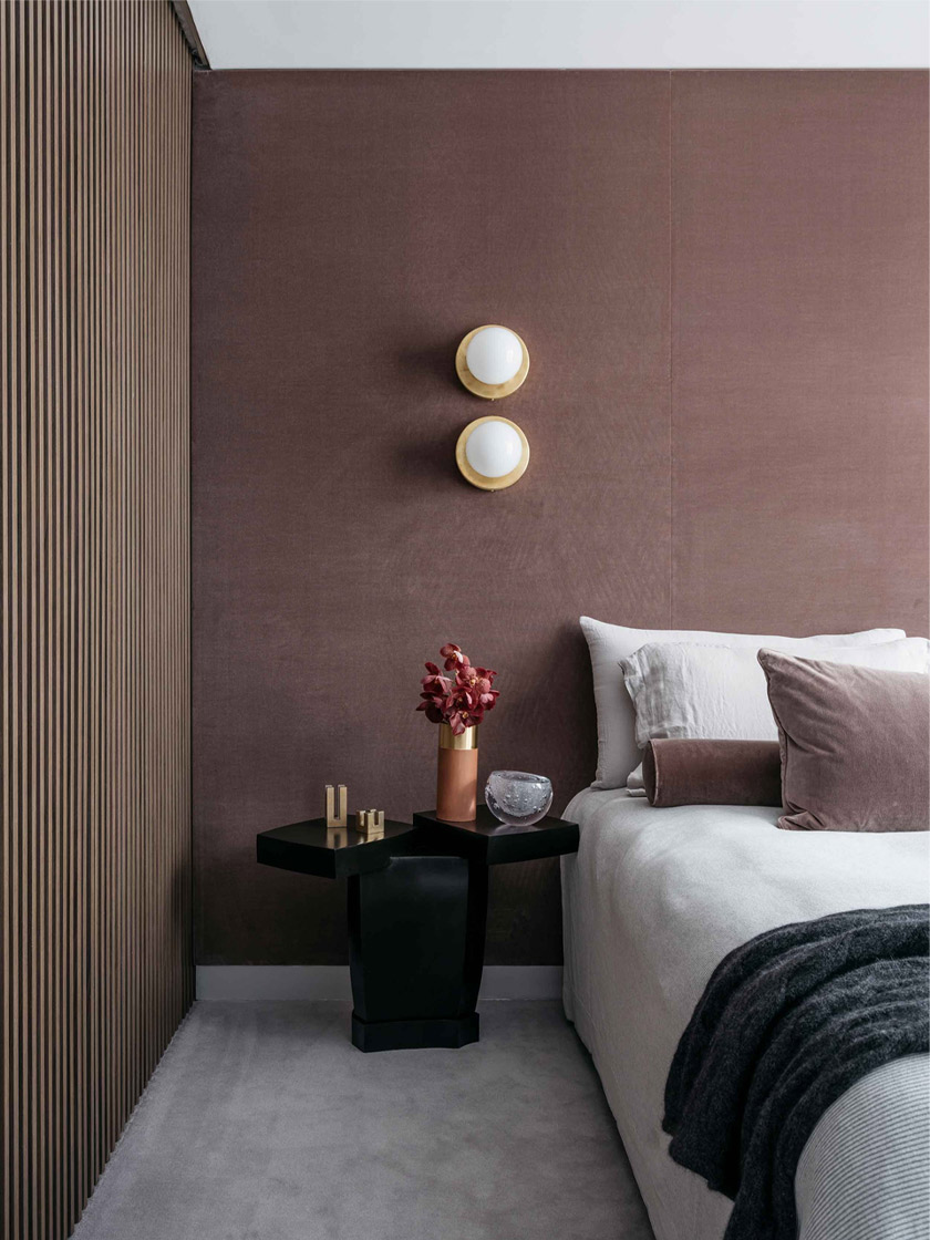 A sophisticated bedroom in The Glasshouse by Nina Maya Interiors, showcasing a black side table, velvet cushions, and warm lighting, accented by wooden wall paneling.