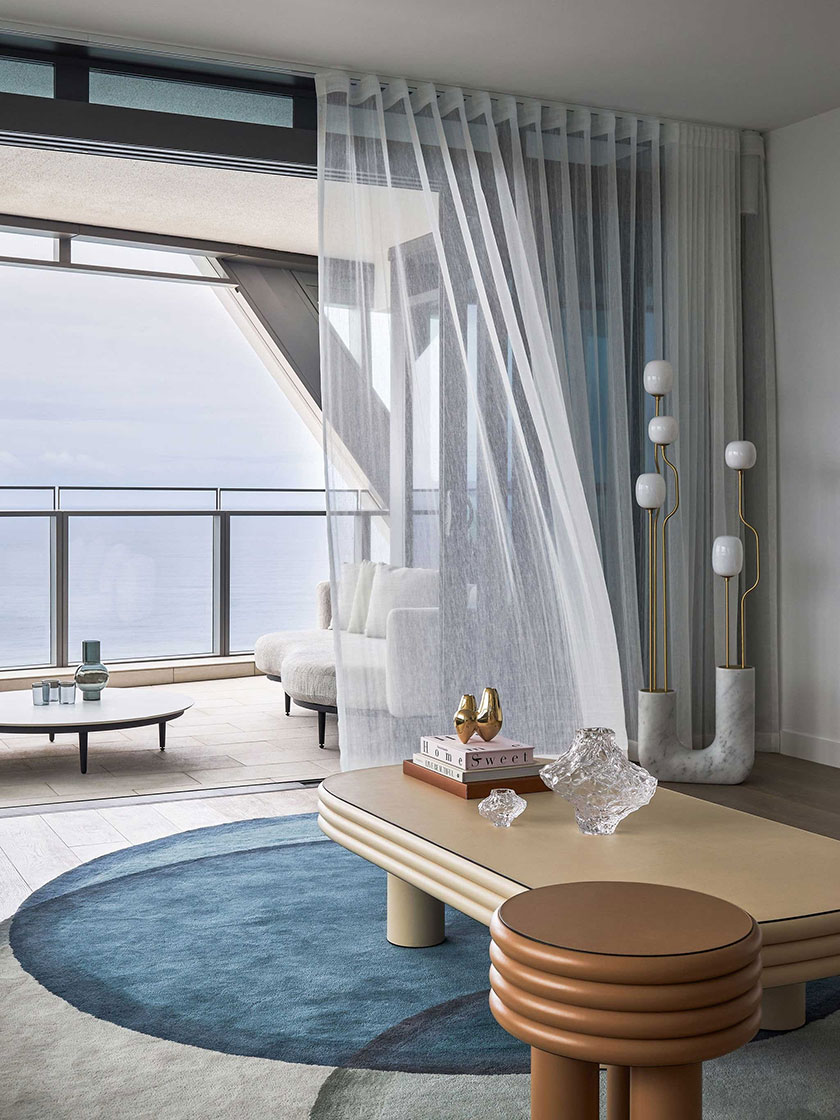 A stylish sitting area designed by Nina Maya Interiors, featuring sheer white curtains, a light beige coffee table with crystal decor, and a view of the balcony with ocean scenery.