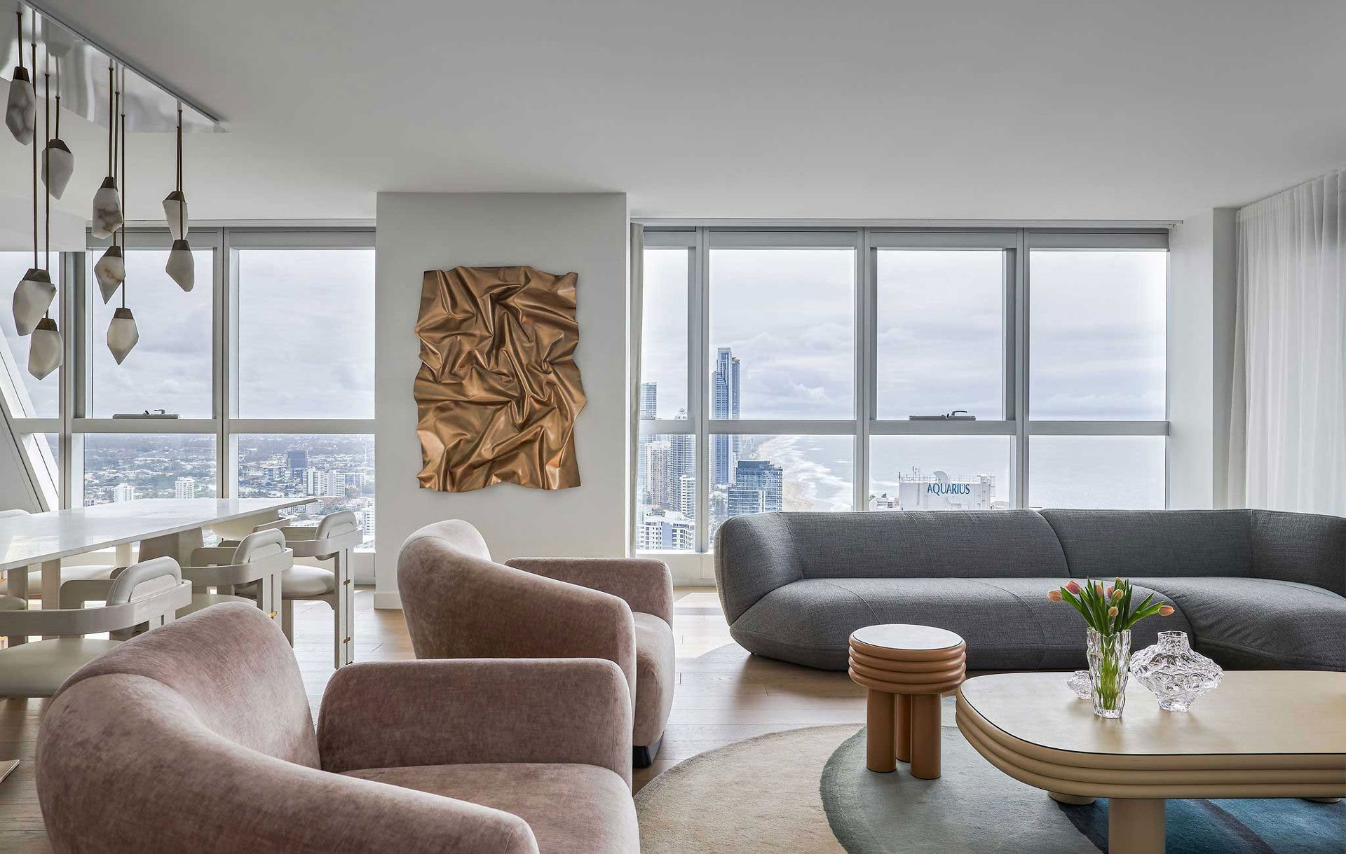 The open-plan living and dining area in the Jewel Apartment by Nina Maya Interiors, showcasing contemporary furniture in muted tones, a bronze wall sculpture, and a panoramic view of the ocean and city skyline.