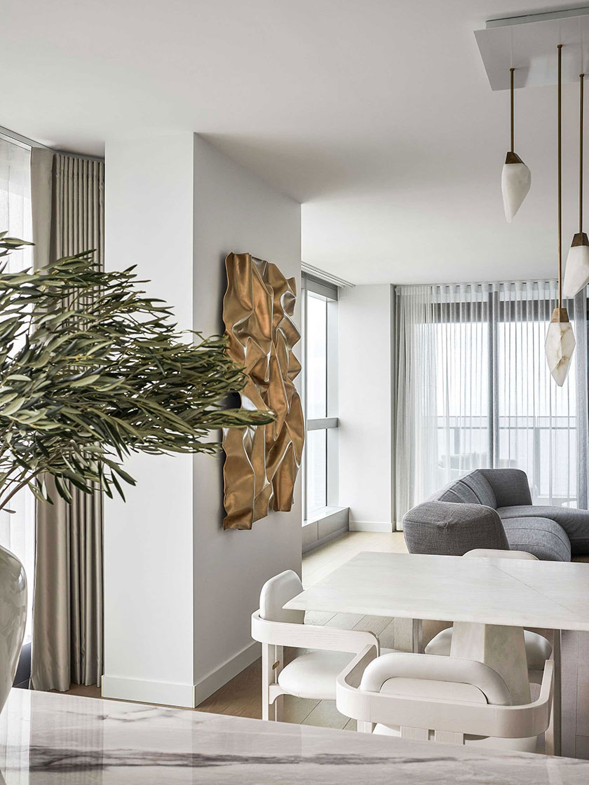 The dining area in the Jewel Apartment by Nina Maya Interiors, showcasing a marble dining table, white chairs, and a unique brass wall sculpture, with an adjacent living space.