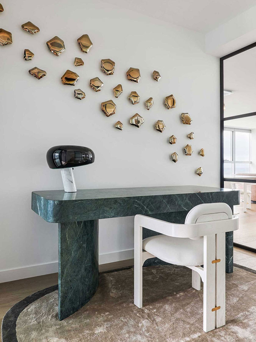 A closer view of the office area in the Jewel Apartment by Nina Maya Interiors, highlighting the green marble desk, white chair, and a modern lamp, with brass wall decorations.