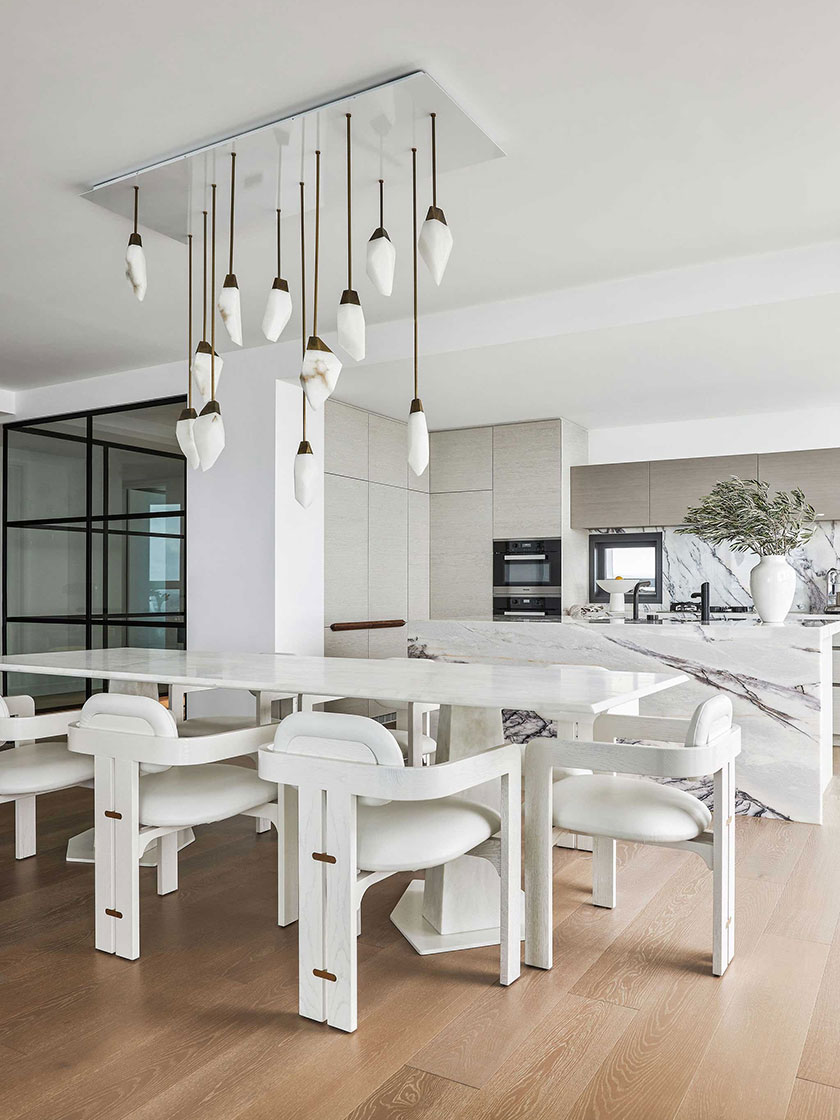 A kitchen and dining space designed by Nina Maya Interiors, with a white marble dining table, white chairs, and a pendant light installation, complemented by modern kitchen decor.