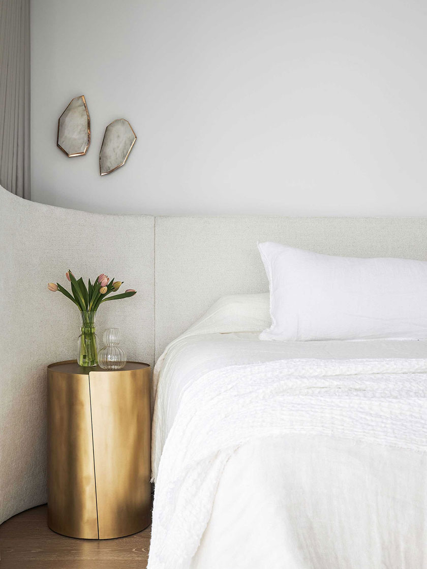 A serene bedroom in the Jewel Apartment by Nina Maya Interiors, featuring a white bed with plush bedding, a gold cylindrical bedside table, and wall-mounted decor.