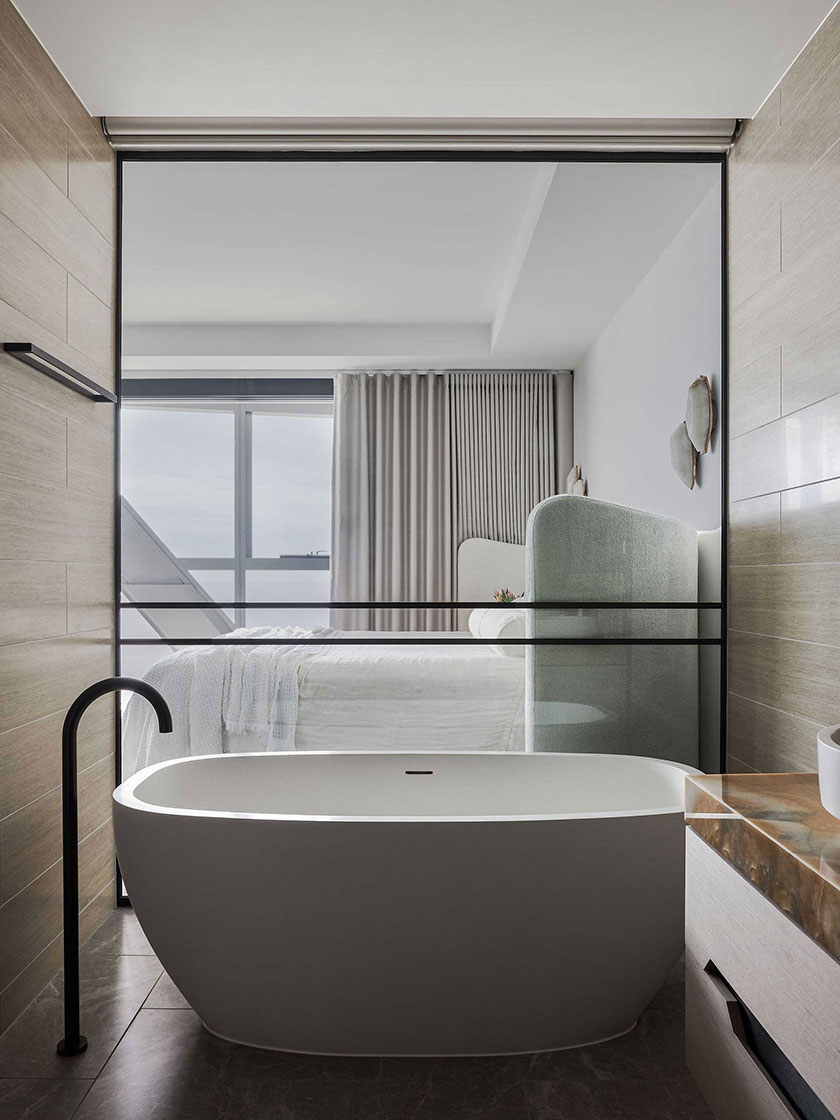 View of the bedroom from the bathroom in the Jewel Apartment by Nina Maya Interiors, showcasing a freestanding bathtub, glass partition, and a cozy bed with soft linens.