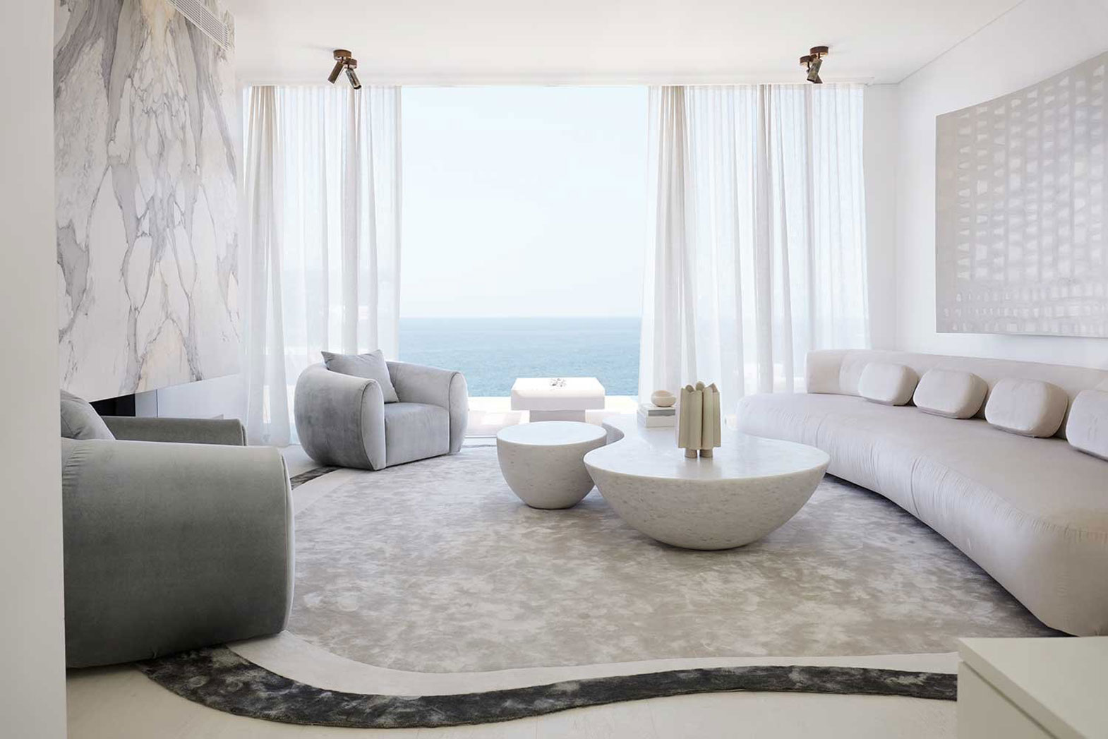 A spacious living room featuring a curved white sofa, grey armchairs, round marble coffee tables, and an ocean view. Interior design by Nina Maya Interiors.