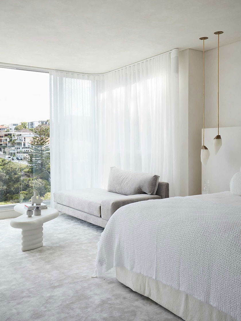 Bright bedroom with white bedding, a grey lounge chair, and an ocean view, designed by Nina Maya Interiors.