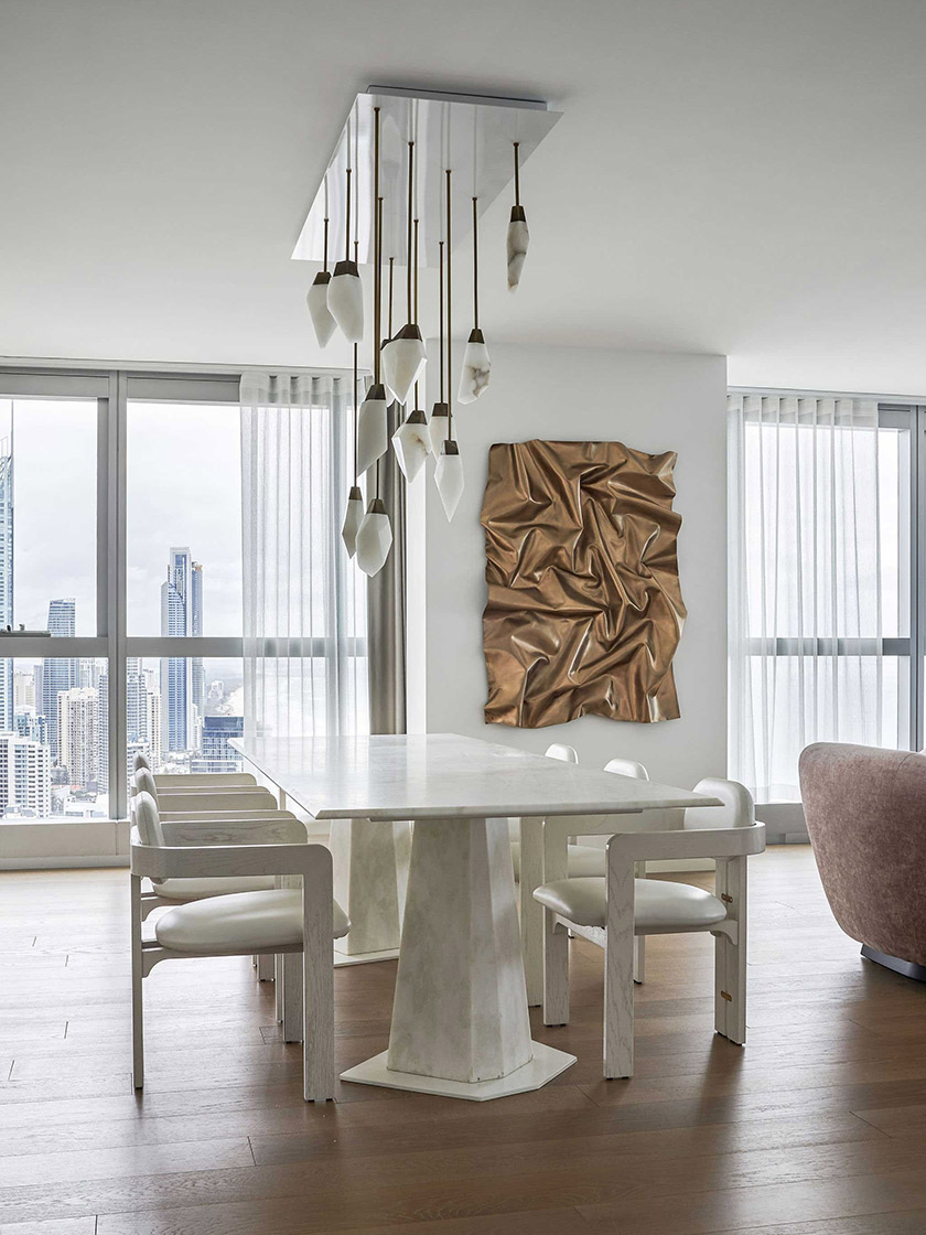 Dining area in the Jewel Apartment by Nina Maya Interiors, featuring a white marble table, white chairs, and a brass and onyx pendant-light installation, with large windows showcasing city views.