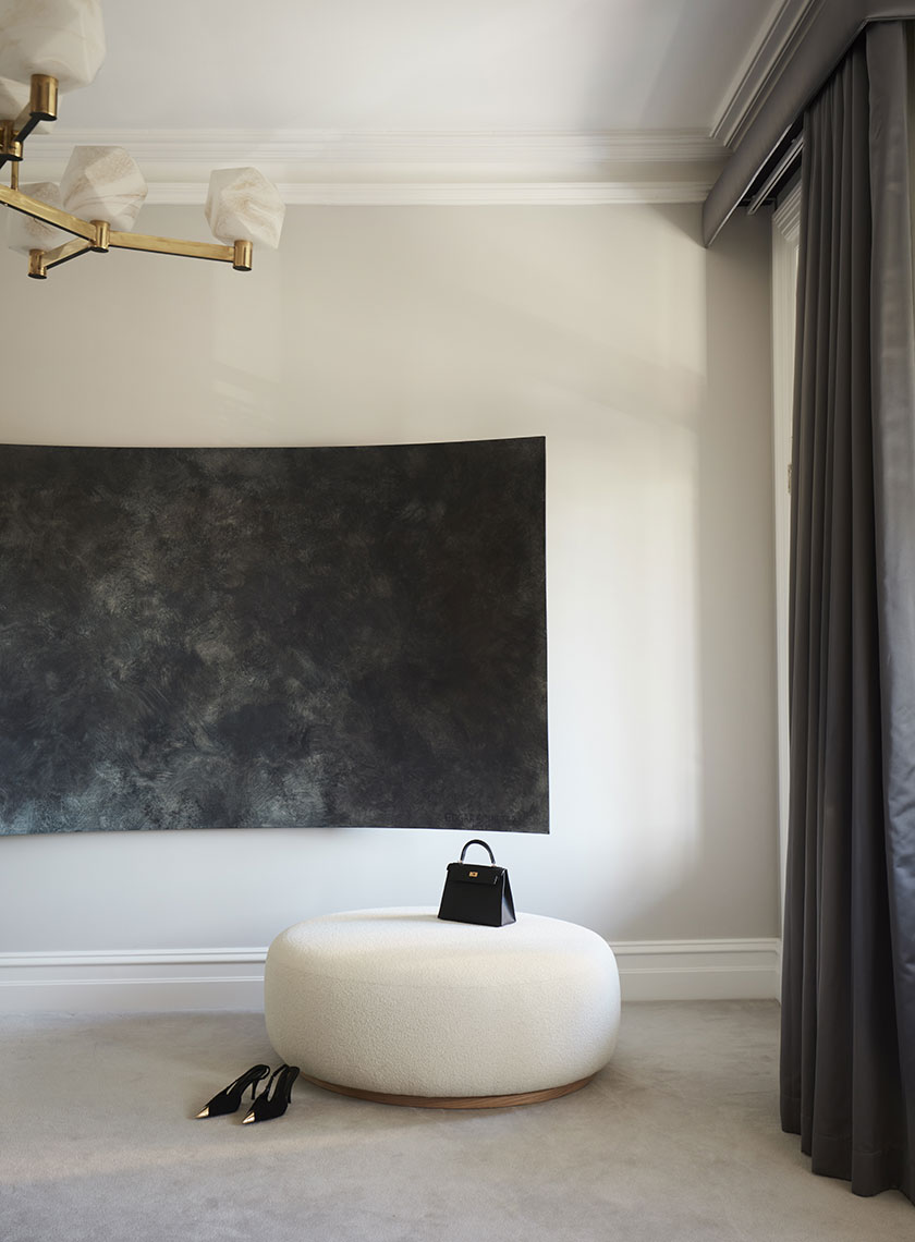 A sophisticated interior shot featuring a black textured wall art piece above a plush, circular white ottoman. A small black handbag rests on the ottoman, while a pair of pointed black heels lie nearby on the carpeted floor, emphasizing minimalist luxury in the design by Nina Maya Interiors. The room is softly lit, with grey curtains partially visible on the right.