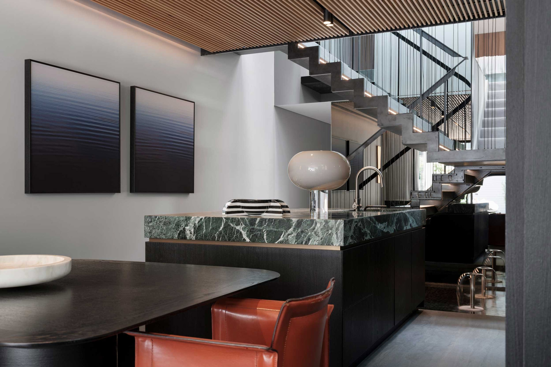 The kitchen and staircase in The Glasshouse by Nina Maya Interiors, featuring green marble countertops, a modern staircase with glass railings, and abstract wall art, blending functionality with style.