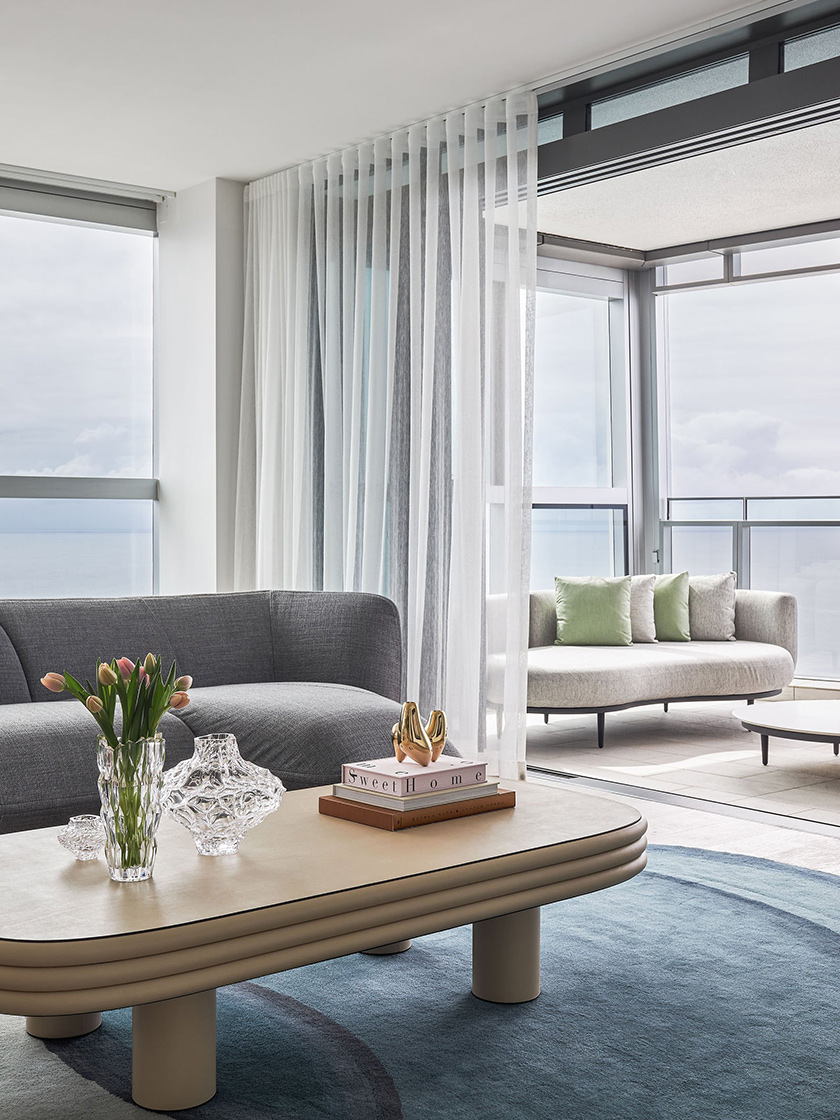 The Jewel Apartment's living area by Nina Maya Interiors, with a gray couch, light beige coffee table, crystal vase, and tulips, complemented by a view of the ocean and balcony seating.