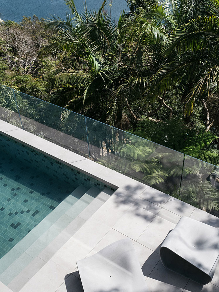 A chic poolside area with minimalist grey lounge chairs and a tiled pool, surrounded by greenery. This outdoor space is part of Nina Maya Interiors' Residence R project, offering a tranquil retreat.