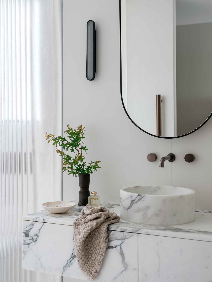 A modern bathroom designed by Nina Maya Interiors featuring a marble countertop and sink, minimalist decor, and a round mirror. The space includes bespoke finishes and a serene, luxurious atmosphere.