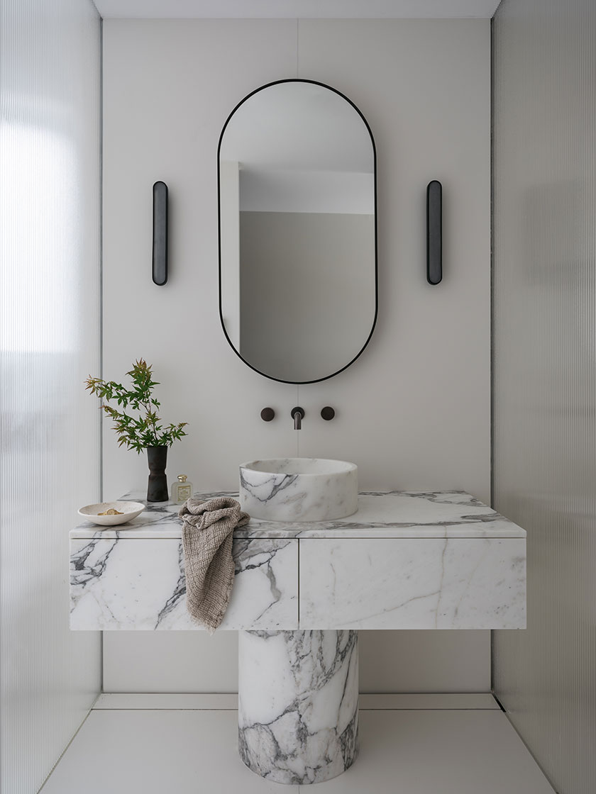 A minimalist bathroom designed by Nina Maya Interiors featuring a marble countertop and cylindrical marble sink base, with an oval mirror and sleek black fixtures.