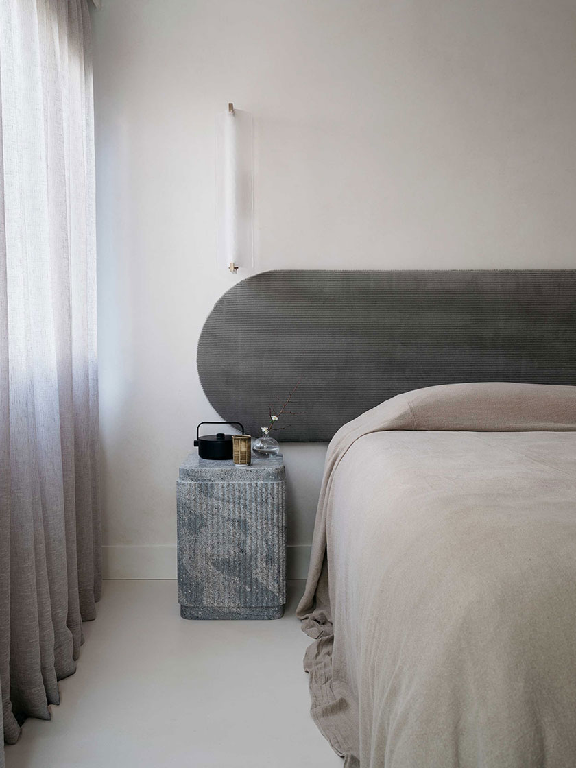 Modern bedroom featuring a grey textured headboard, beige bedding, and a minimalist nightstand, designed by Nina Maya Interiors.