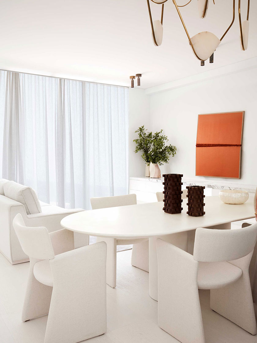 Bright dining area by Nina Maya Interiors, featuring a white oval table, beige chairs, modern chandelier, and light-filled curtains, complemented by an orange abstract painting.