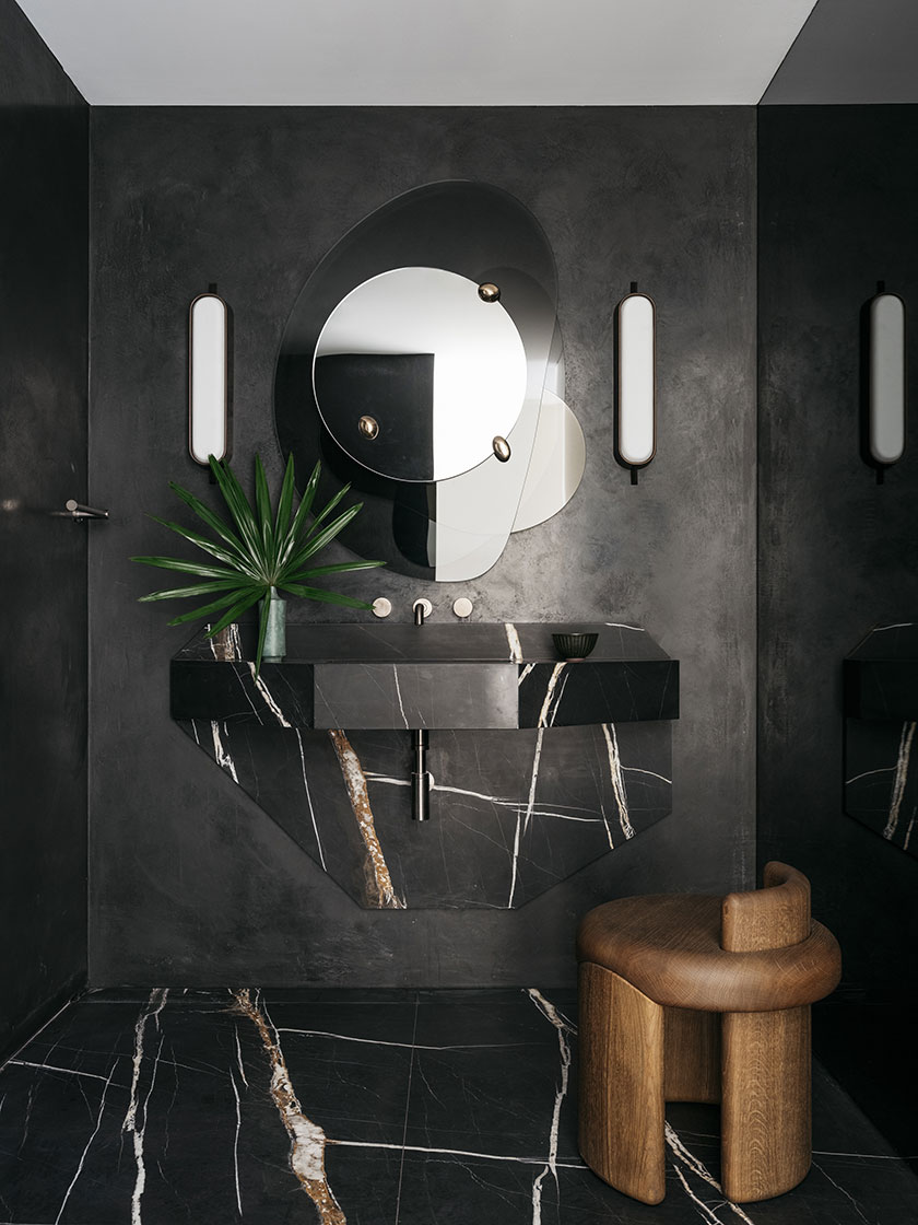 A dramatic powder room with Black Tiger Marble surfaces and a unique, layered mirror design, accented by a small wooden stool. Design by Nina Maya Interiors.