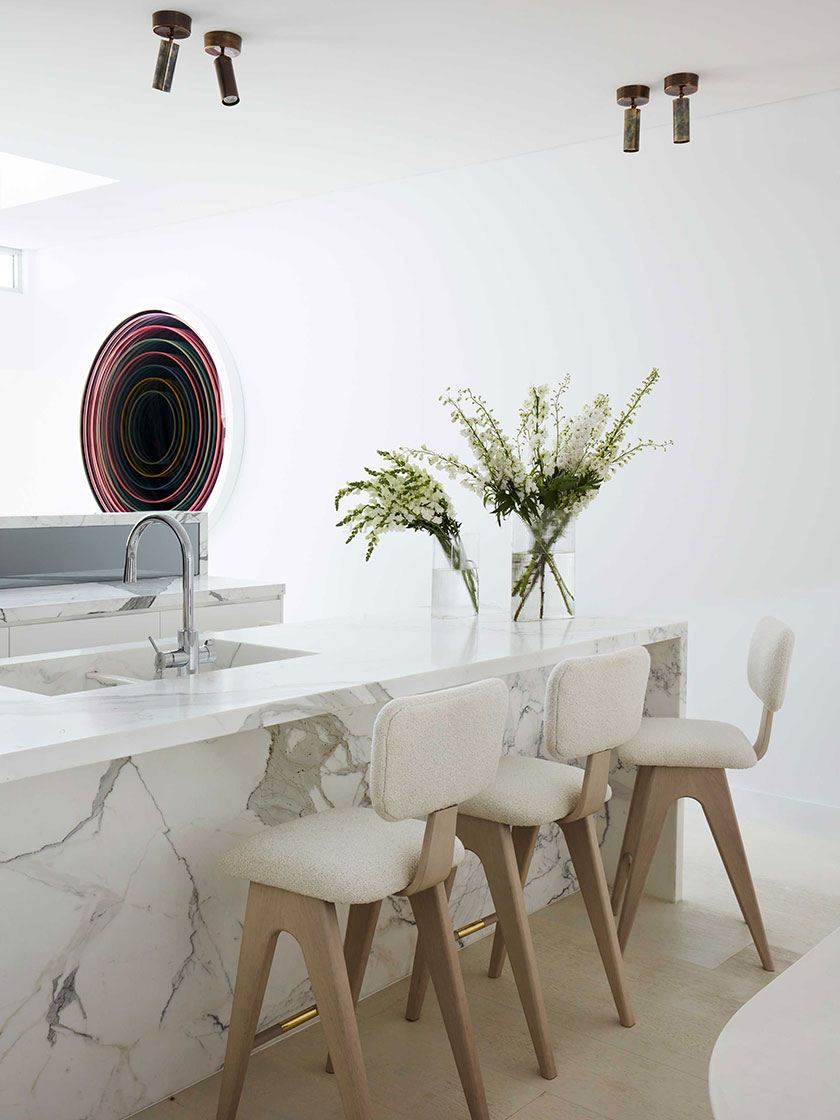 Bright, modern kitchen designed by Nina Maya Interiors, with a marble island, beige stools, and white floral arrangements, accompanied by a colorful circular artwork on the wall.