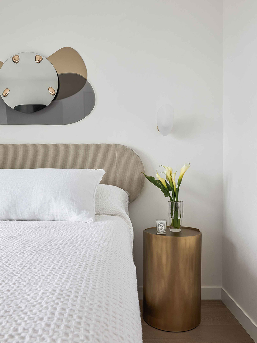 Bedroom in the Jewel Apartment by Nina Maya Interiors, featuring a beige upholstered headboard, white bedding, a gold bedside table, and minimalist wall decor.