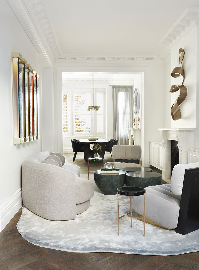 A sophisticated living room with curved sofas, green marble coffee tables, and sculptural bronze wall art, leading into a bright dining area with large windows. Interior design by Nina Maya Interiors.