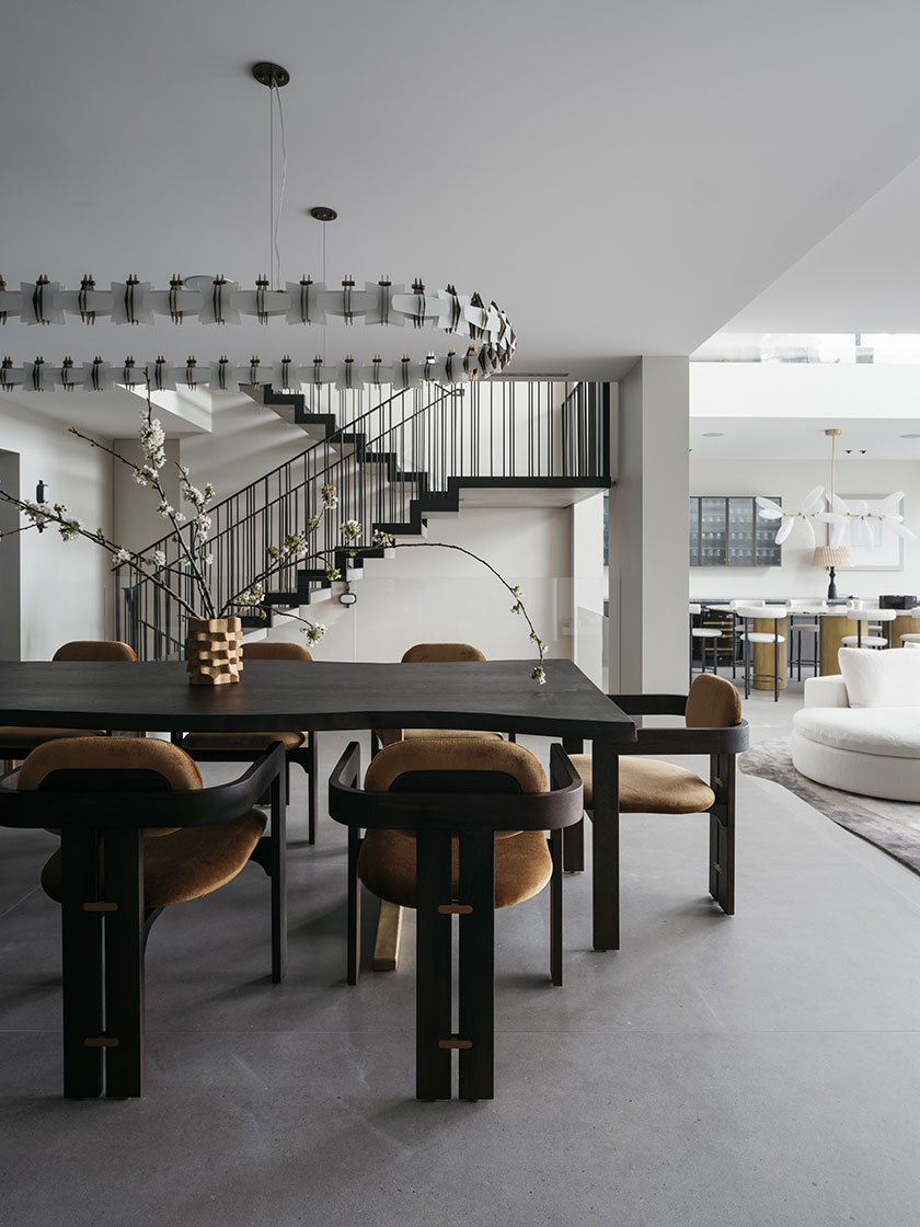 An open-plan dining area designed by Nina Maya Interiors, featuring a dark wood dining table, custom chairs, and a sculptural chandelier, with a view of a floating staircase in the background.