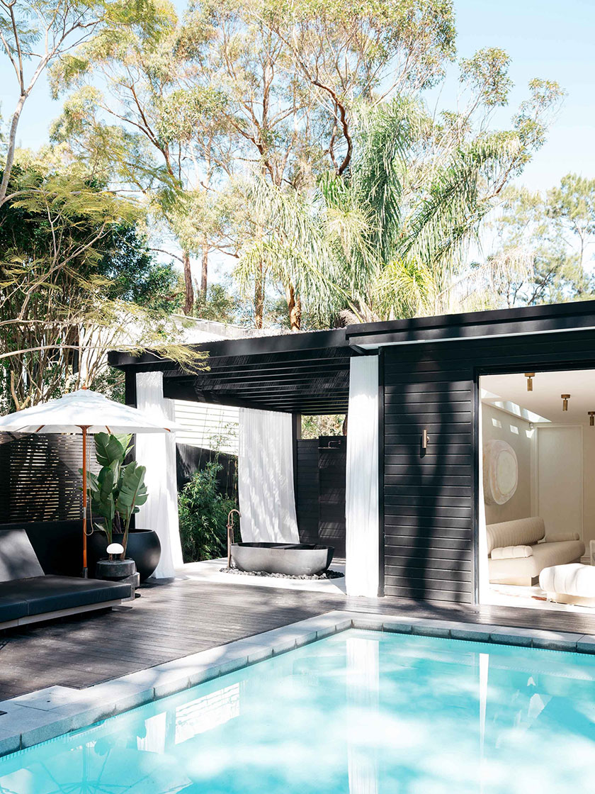 Elegant poolside lounge with black furniture, a white umbrella, and lush greenery, designed by Nina Maya Interiors.