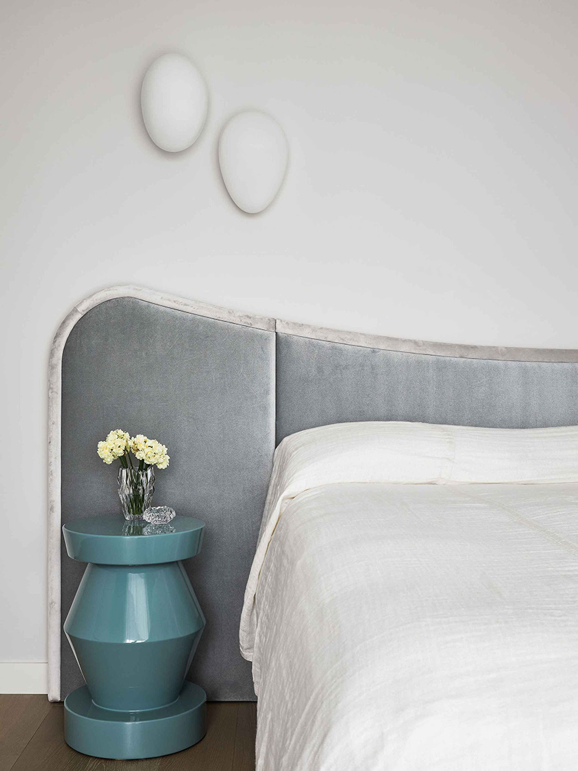 A serene bedroom in the Jewel Apartment by Nina Maya Interiors, featuring a soft gray upholstered headboard, white bedding, and a teal bedside table with fresh flowers, accented by white wall-mounted decor.