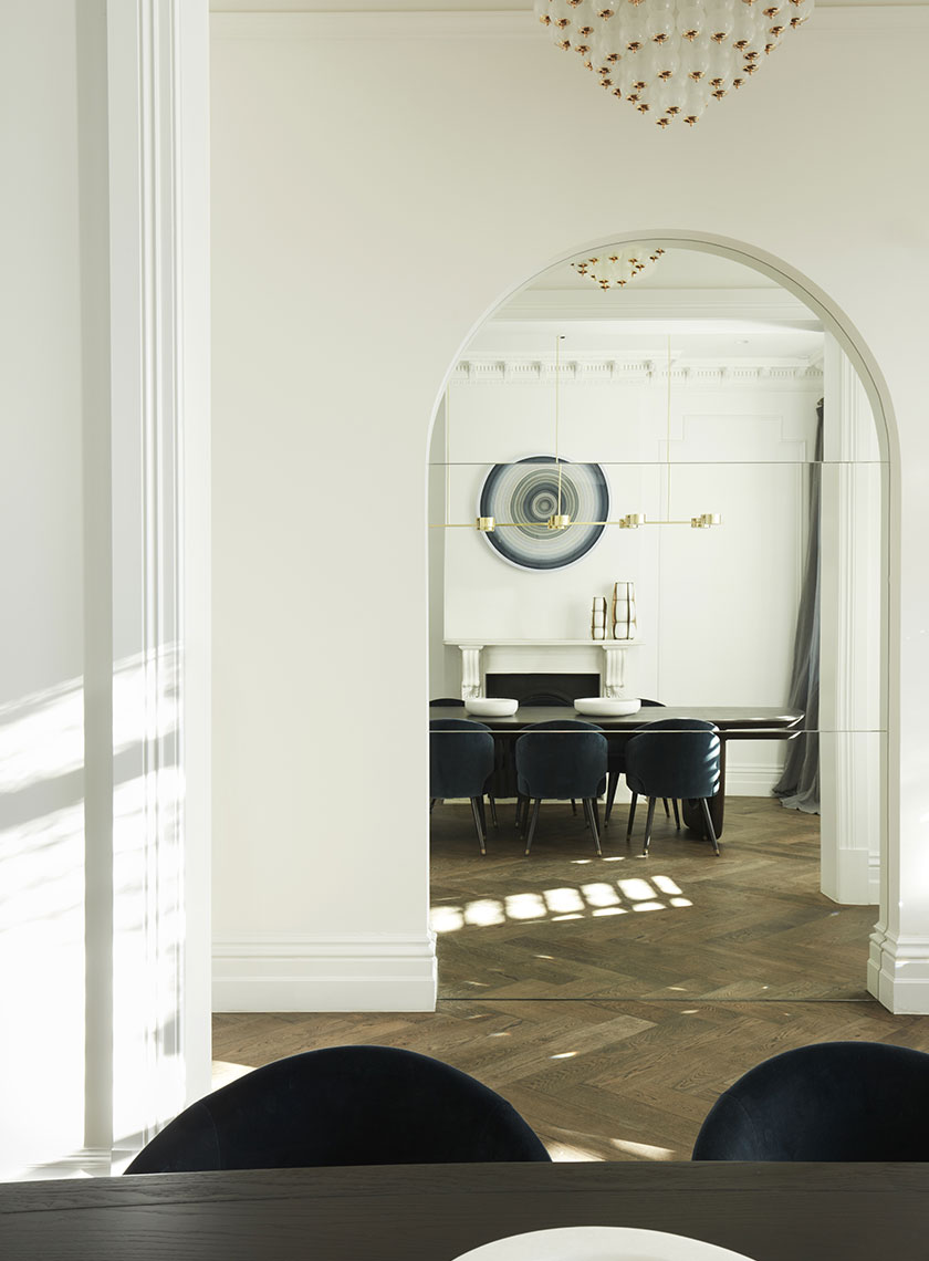 A formal dining room viewed through a large archway, featuring a modern brass chandelier, dark wood dining table, and a circular piece of art on the wall. Interior design by Nina Maya Interiors.