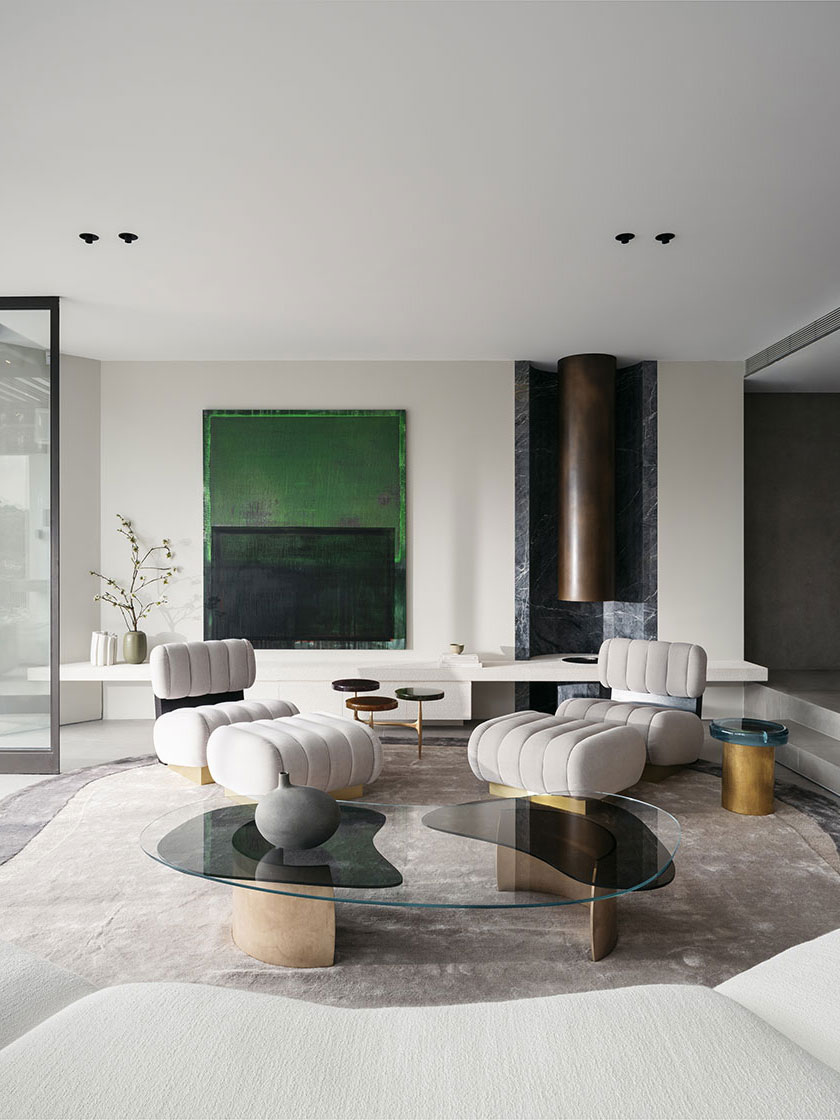 A modern living room designed by Nina Maya Interiors with plush grey chairs, a unique glass coffee table, and a large green painting. The room is accented with brass and marble elements.