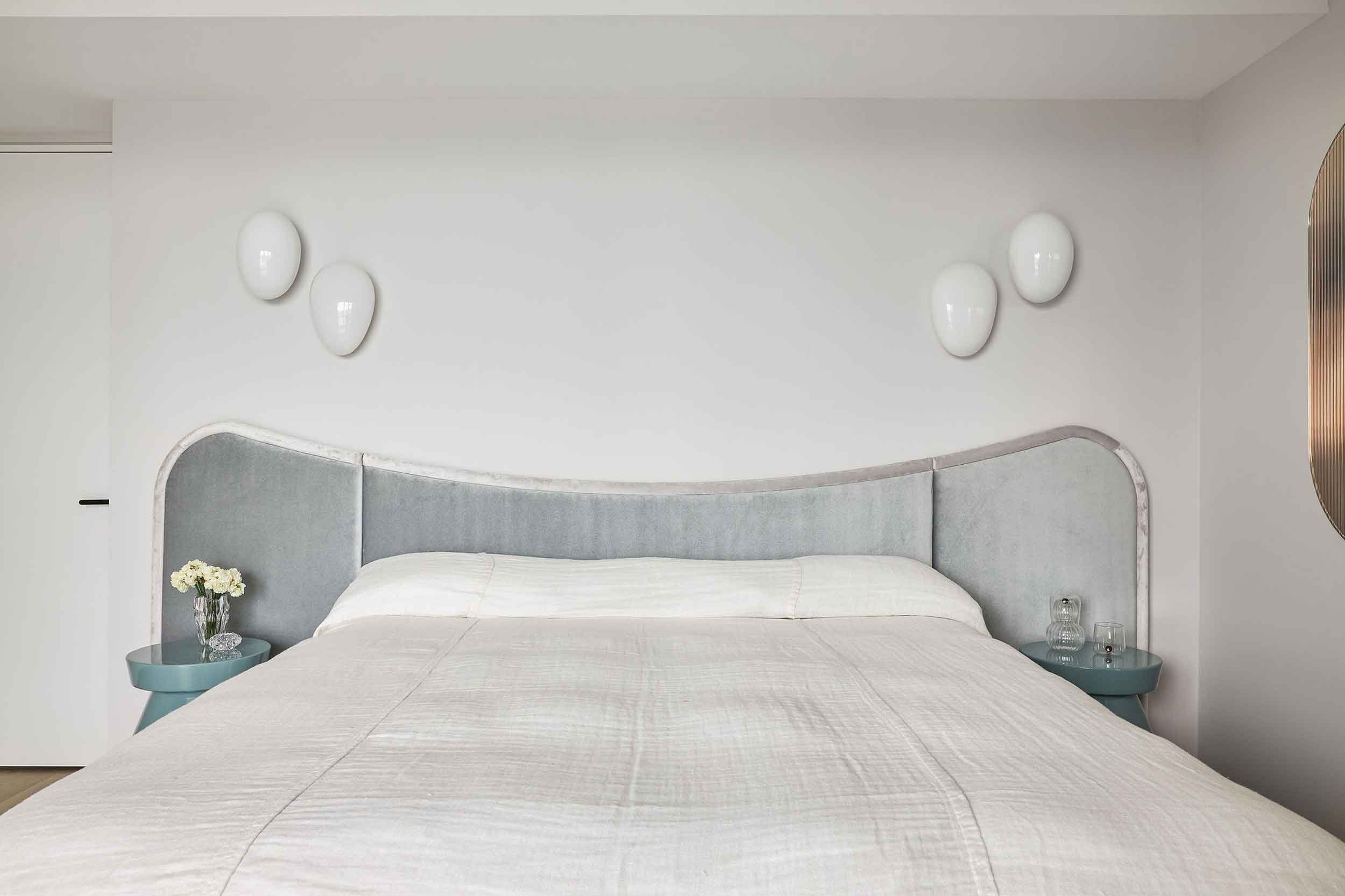 A full view of the bedroom in the Jewel Apartment by Nina Maya Interiors, showcasing a gray upholstered headboard, white bedding, teal bedside tables, and minimalist white wall decor.