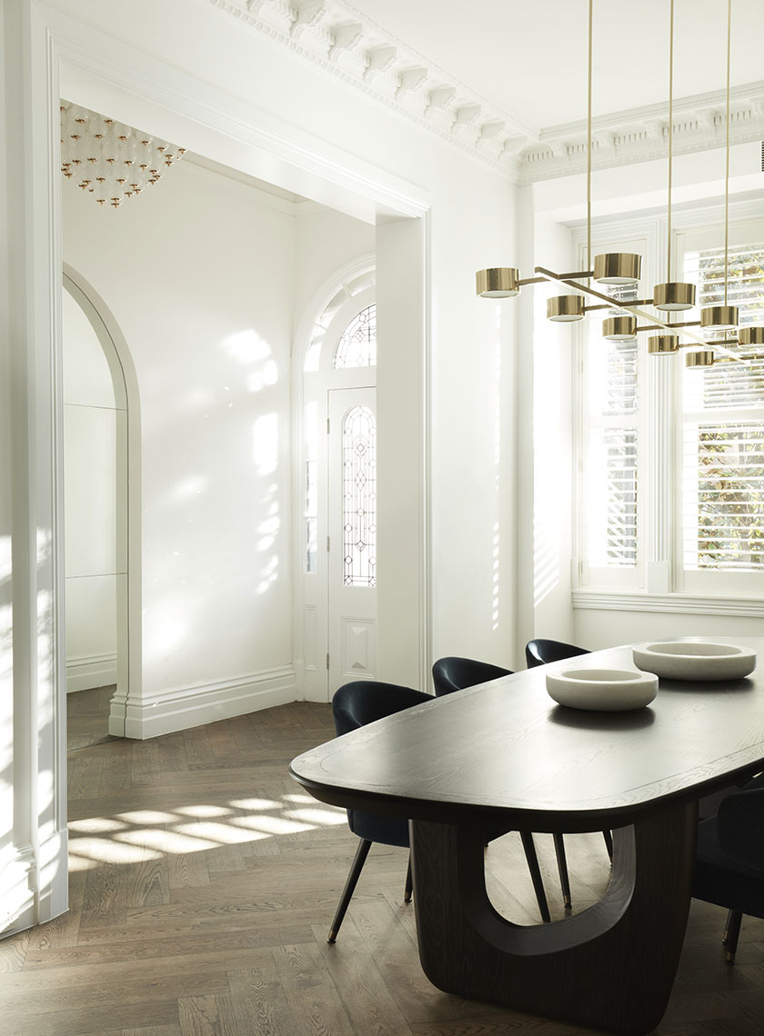 A dining room with a large, dark wood table surrounded by blue velvet chairs, illuminated by a contemporary brass chandelier under a white, detailed ceiling. Interior design by Nina Maya Interiors.