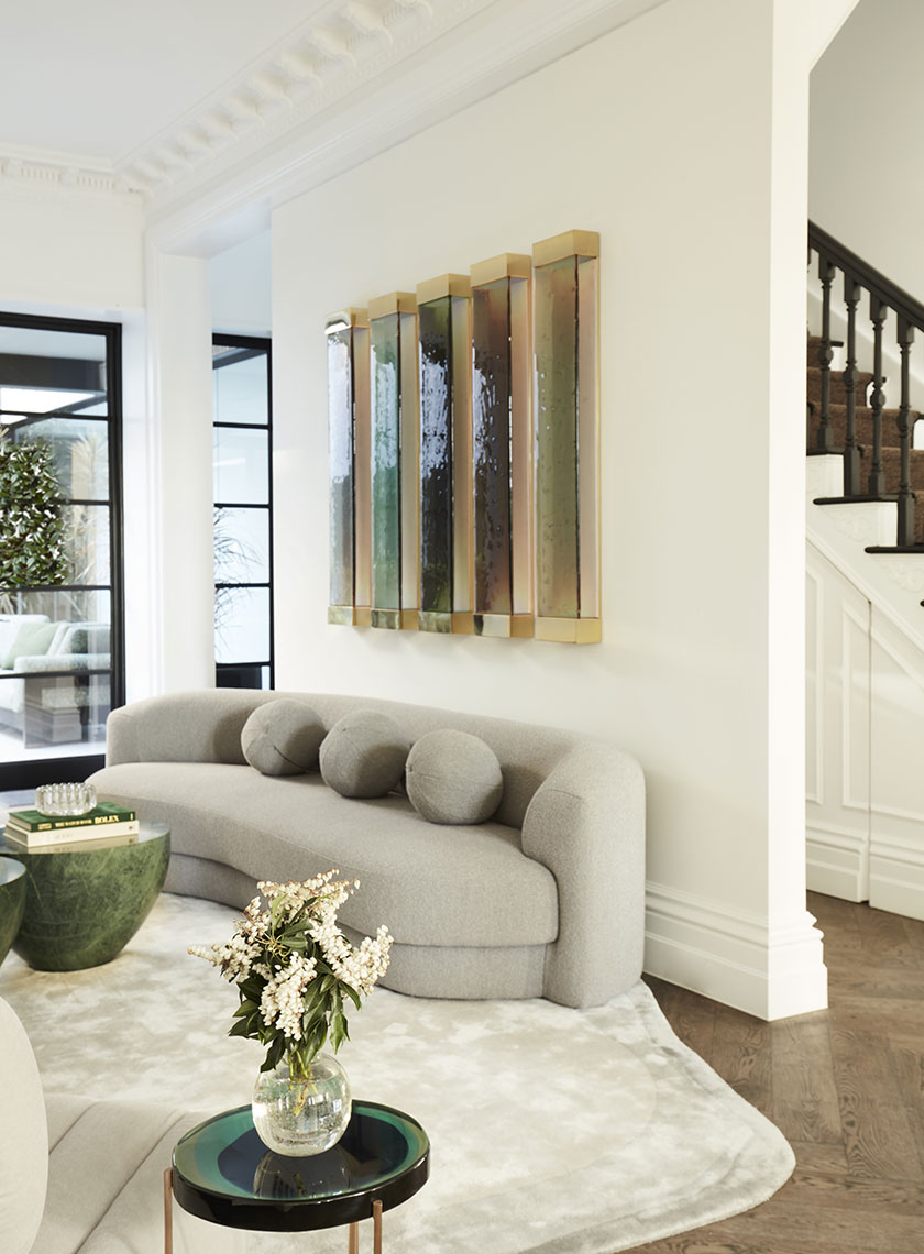 A close-up of a curved gray sofa with round cushions, positioned in front of modern, backlit wall art in warm tones. Interior design by Nina Maya Interiors.