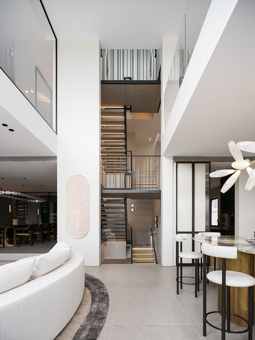 A modern, open-plan living area designed by Nina Maya Interiors, featuring a view of a floating staircase with bronze and porcelain steps that runs along the entire axis of the building. The space includes a white curved sofa and a marble kitchen island with white stools. The minimalist decor emphasizes natural light and clean lines.