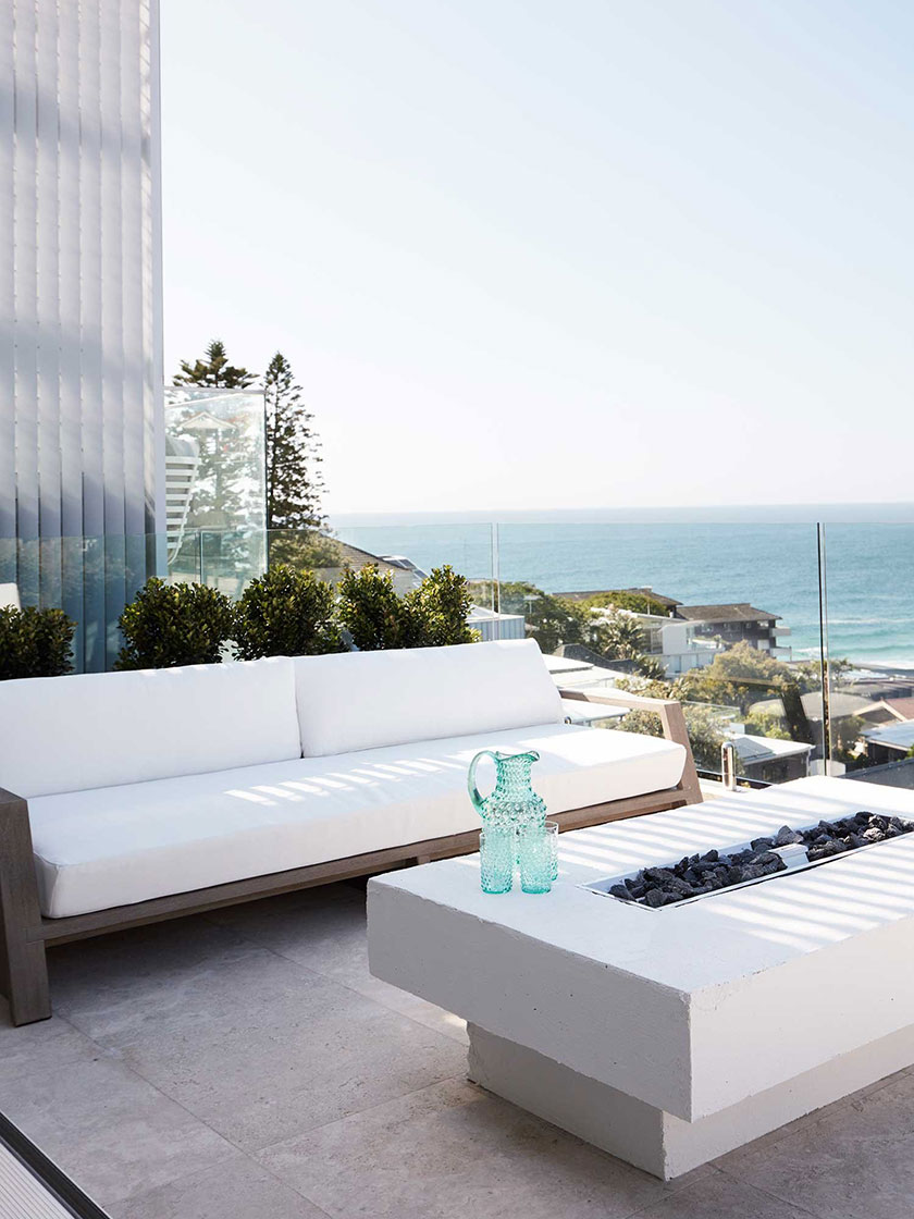 Modern outdoor seating area with white cushions and a fire pit, overlooking the ocean.