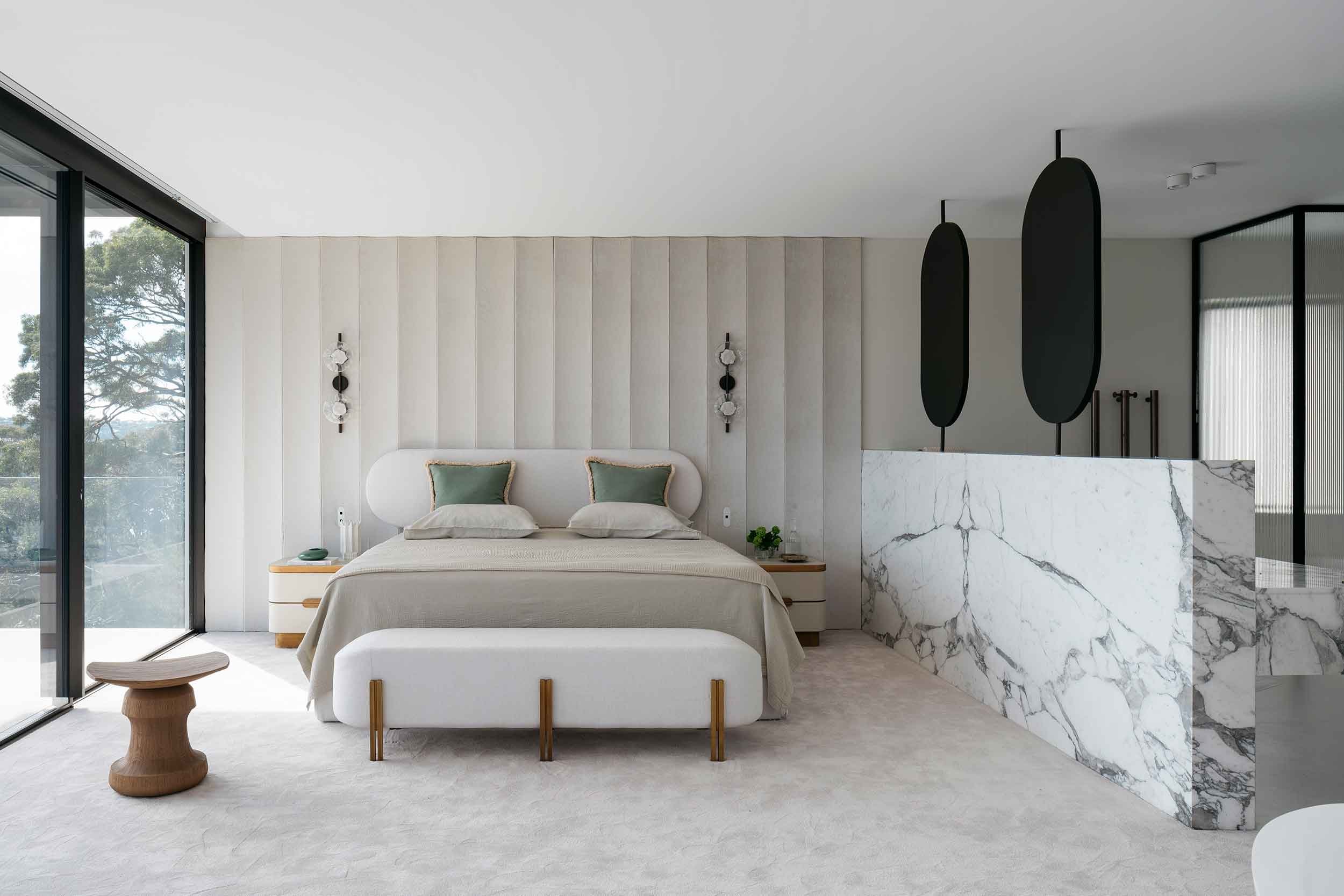 A luxurious bedroom designed by Nina Maya Interiors featuring a white upholstered bed with green accent pillows, a marble partition, and large glass doors offering a view of the outdoors. The room includes modern pendant lights and custom bedside tables.