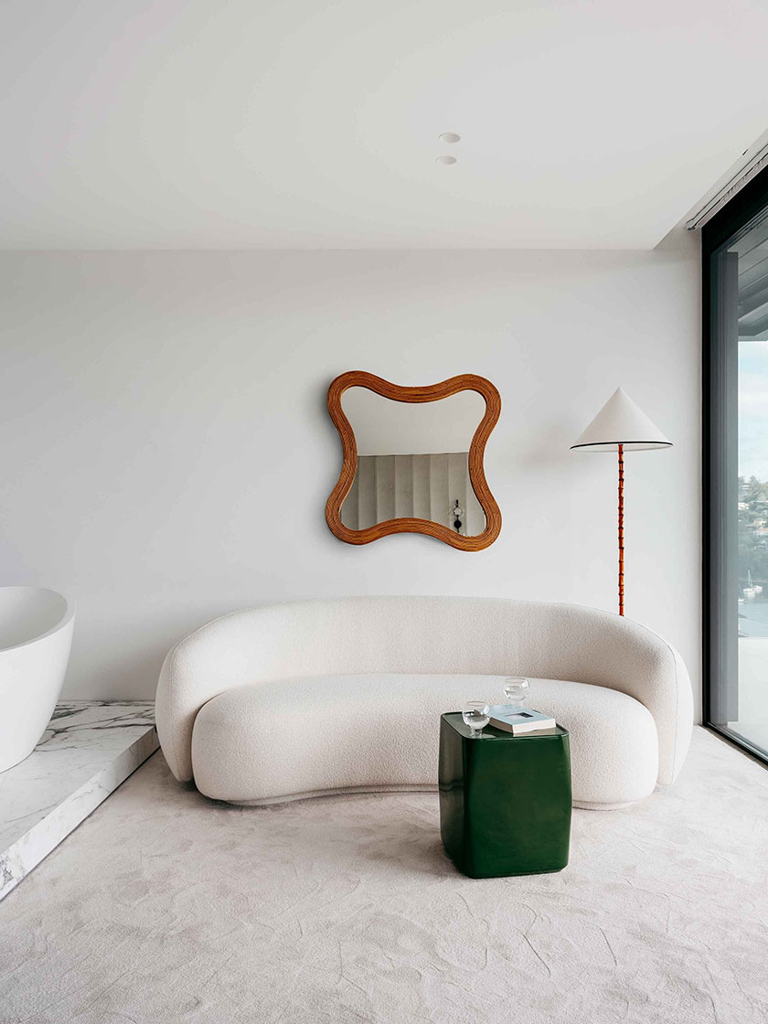 A cozy seating nook in the bedroom with a curved white sofa, a green side table, and a large mirror. Interior design by Nina Maya Interiors.