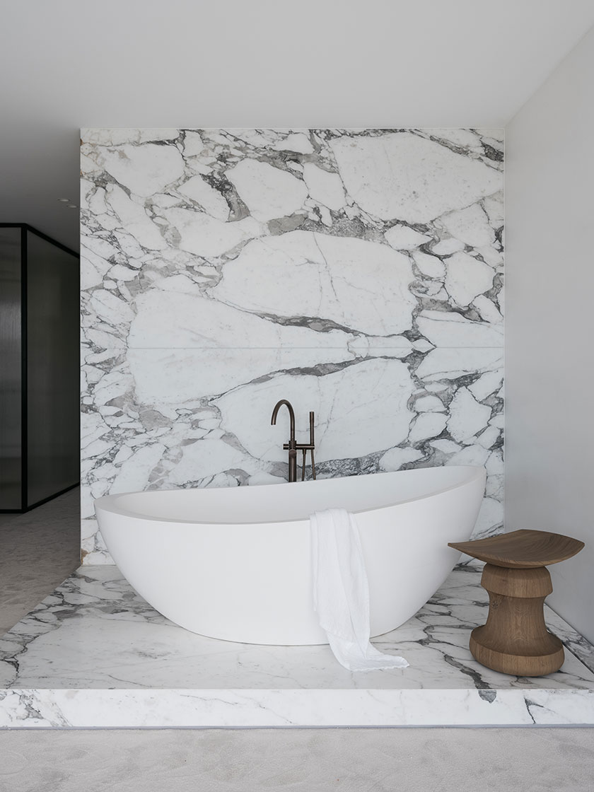 A luxurious bathroom designed by Nina Maya Interiors featuring a freestanding white bathtub on a marble platform, set against a striking marble wall. A wooden stool and a white towel add to the serene and elegant atmosphere.