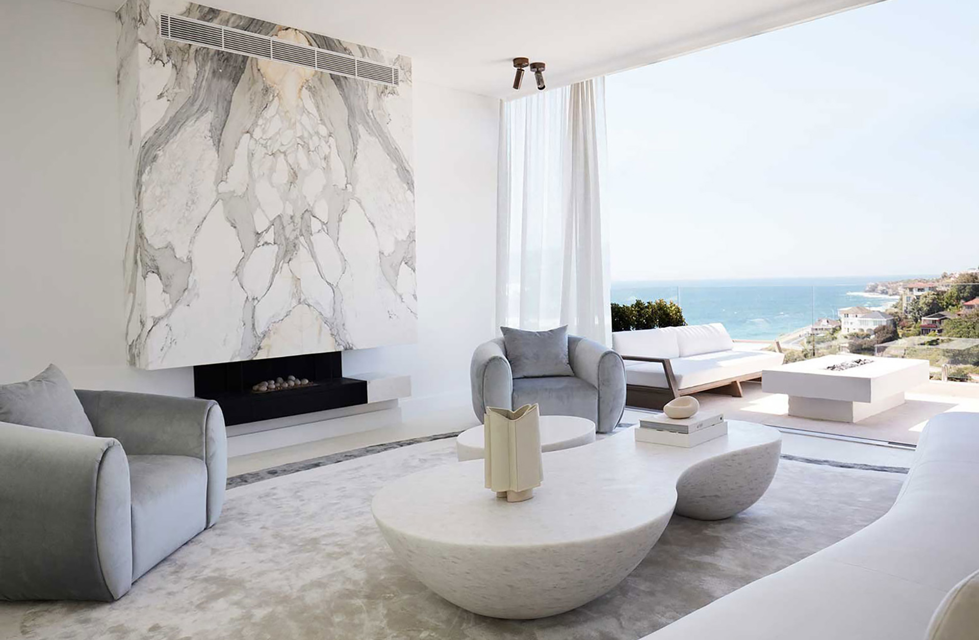 A bright living room with a curved white sofa, grey armchairs, marble fireplace, and ocean view. Interior design by Nina Maya Interiors