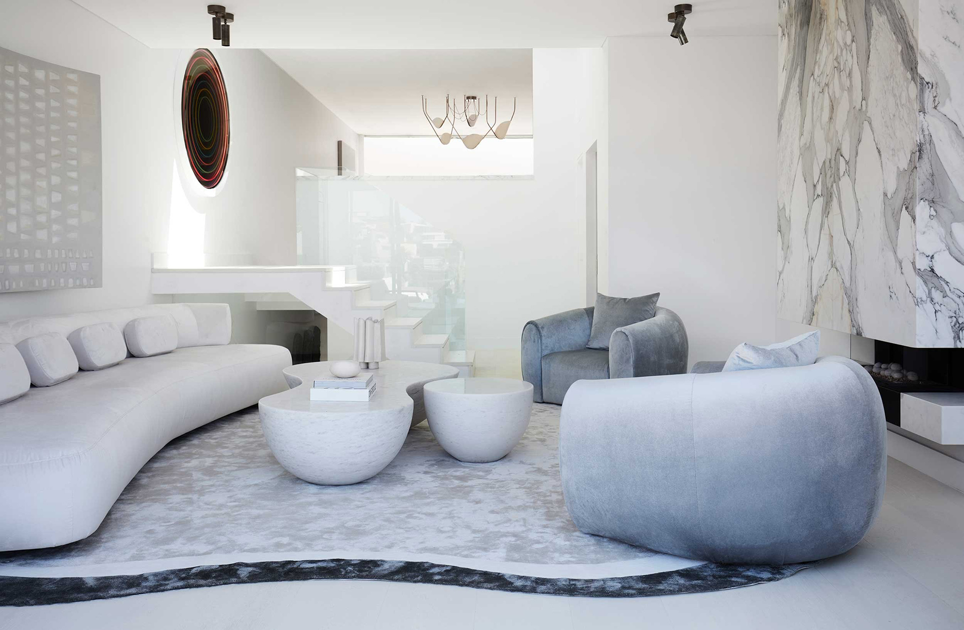 A contemporary living room with a curved white sofa, abstract wall art, marble coffee tables, and a marble fireplace. Interior design by Nina Maya Interiors.