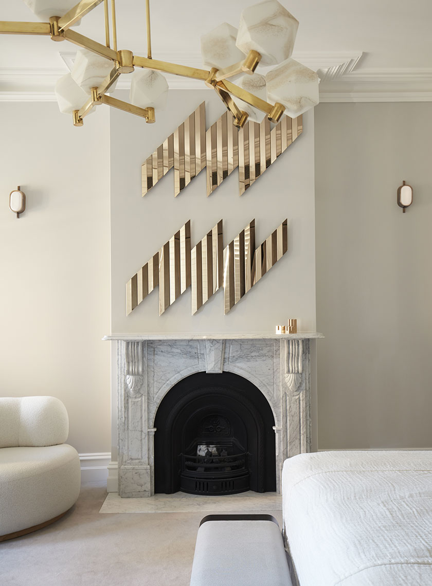 A chic fireplace in a modern room, with a marble surround and a black fireplace insert, accented by gold, geometric wall art above. A brass chandelier with hexagonal lights hangs from the ceiling, complementing the overall luxe aesthetic crafted by Nina Maya Interiors. The room is designed with soft, neutral tones, featuring a curved white sofa in the corner.