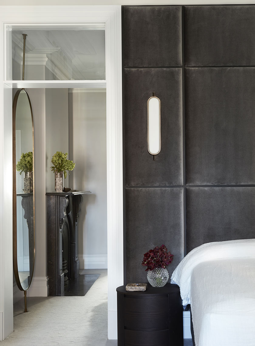 A luxurious walk-in wardrobe with dark, glass-fronted wardrobes illuminated by soft interior lighting. A circular grey ottoman with a black handbag sits in the center, beneath a glass-topped side table. The wardrobe opens into a bright room with a marble fireplace, highlighting the seamless blend of functionality and style by Nina Maya Interiors.