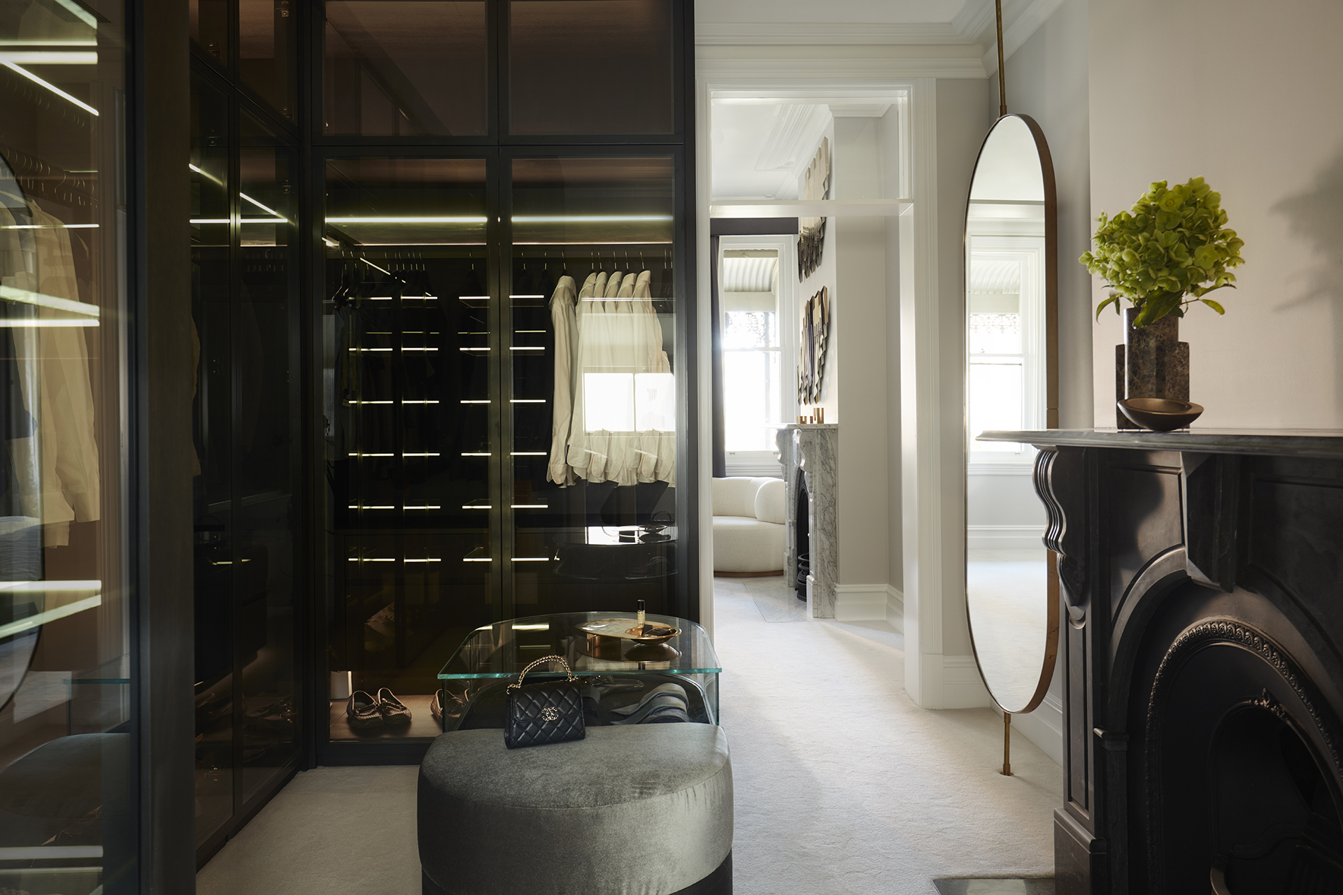 Walk-in Closet with Glass Doors: A luxurious walk-in closet featuring glass-paneled wardrobe doors, a plush round ottoman, and a sleek black fireplace, designed by Nina Maya Interiors at Residence K, Potts Point.