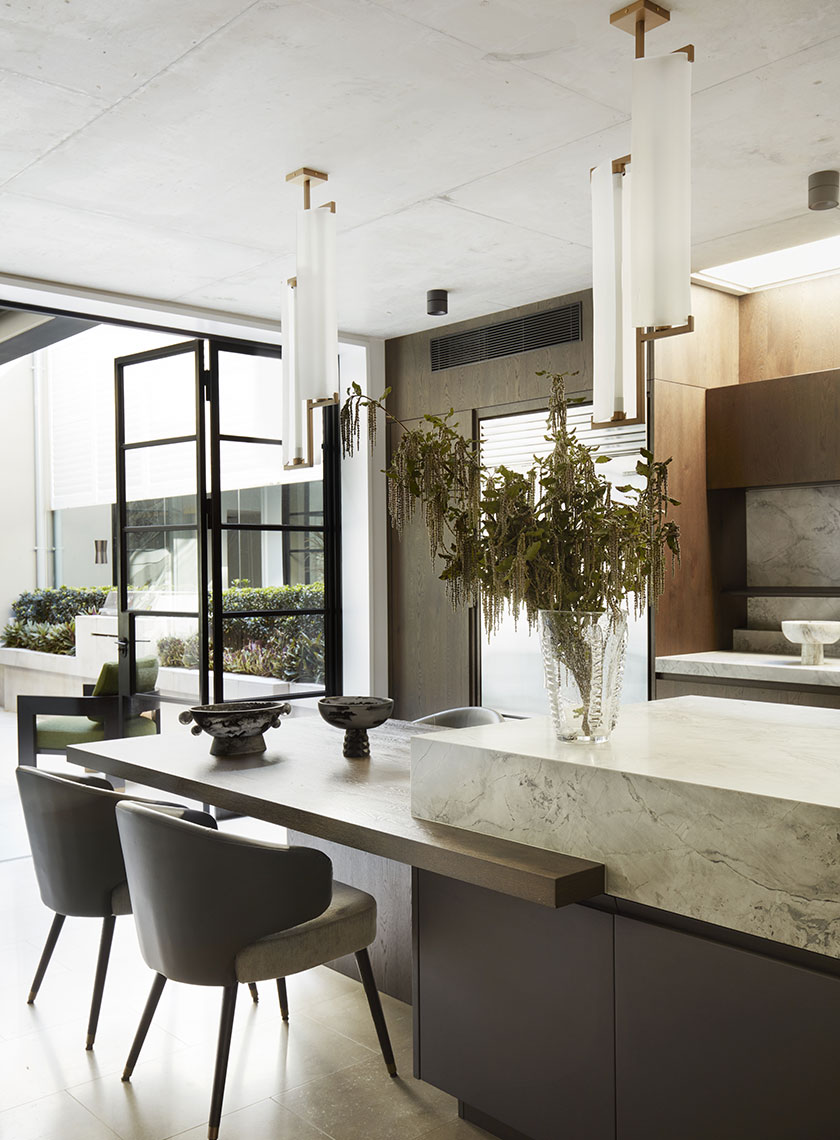 A modern kitchen and dining area featuring a marble island, wooden cabinetry, and a vase of dried branches on the table, with floor-to-ceiling glass doors opening to an outdoor space. Interior design by Nina Maya Interiors.