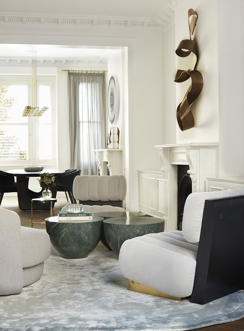 A luxurious living room with plush, light gray armchairs and green marble coffee tables, complemented by sculptural wall art and a soft gray area rug. Interior design by Nina Maya Interiors.