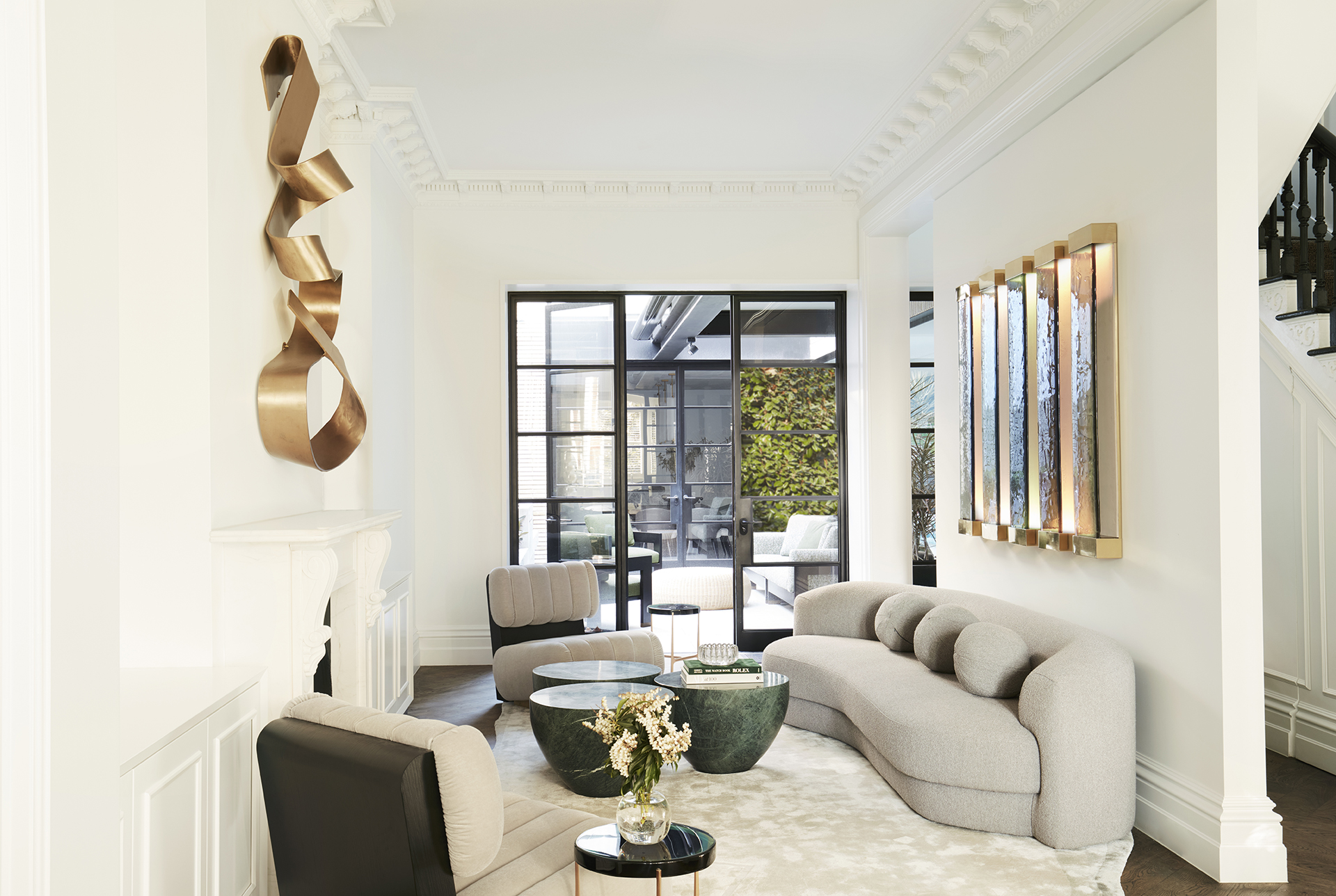 A stylish living room with a curved gray sofa, green marble coffee tables, and modern art on the walls, with a view of an outdoor seating area through glass doors. Interior design by Nina Maya Interiors.