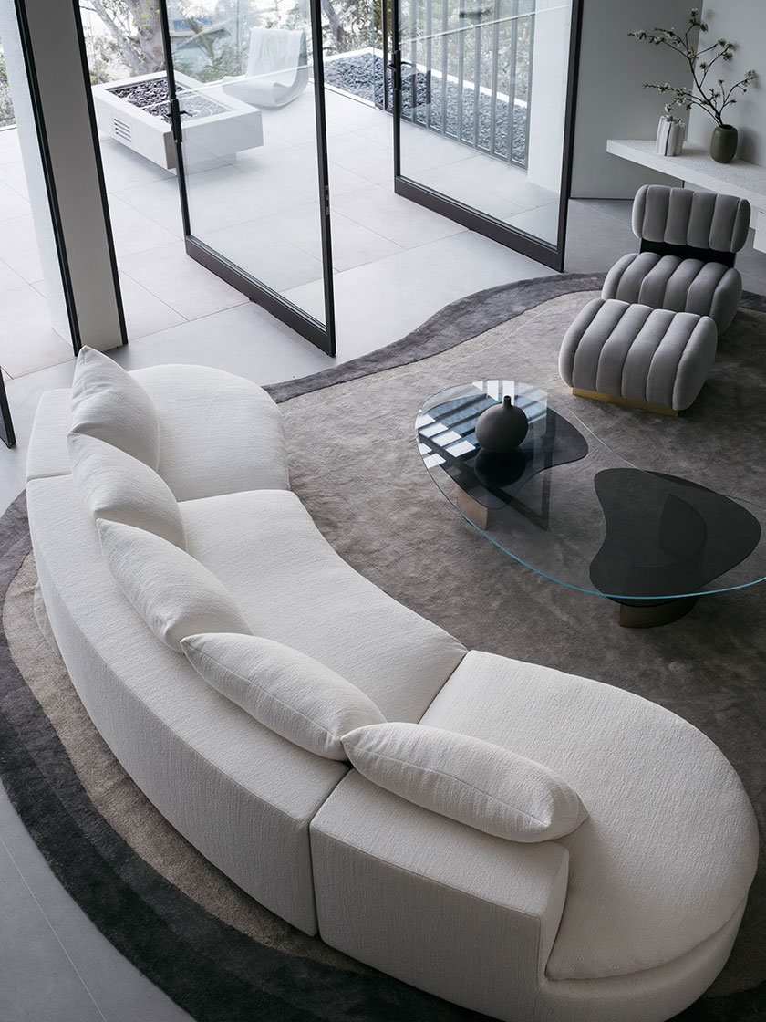 Overhead view of a curved white bouclé sofa with multiple cushions, adjacent to grey upholstered chairs and a glass coffee table on a grey rug. This setup is part of Nina Maya Interiors' design for Residence R, emphasising minimalist luxury.