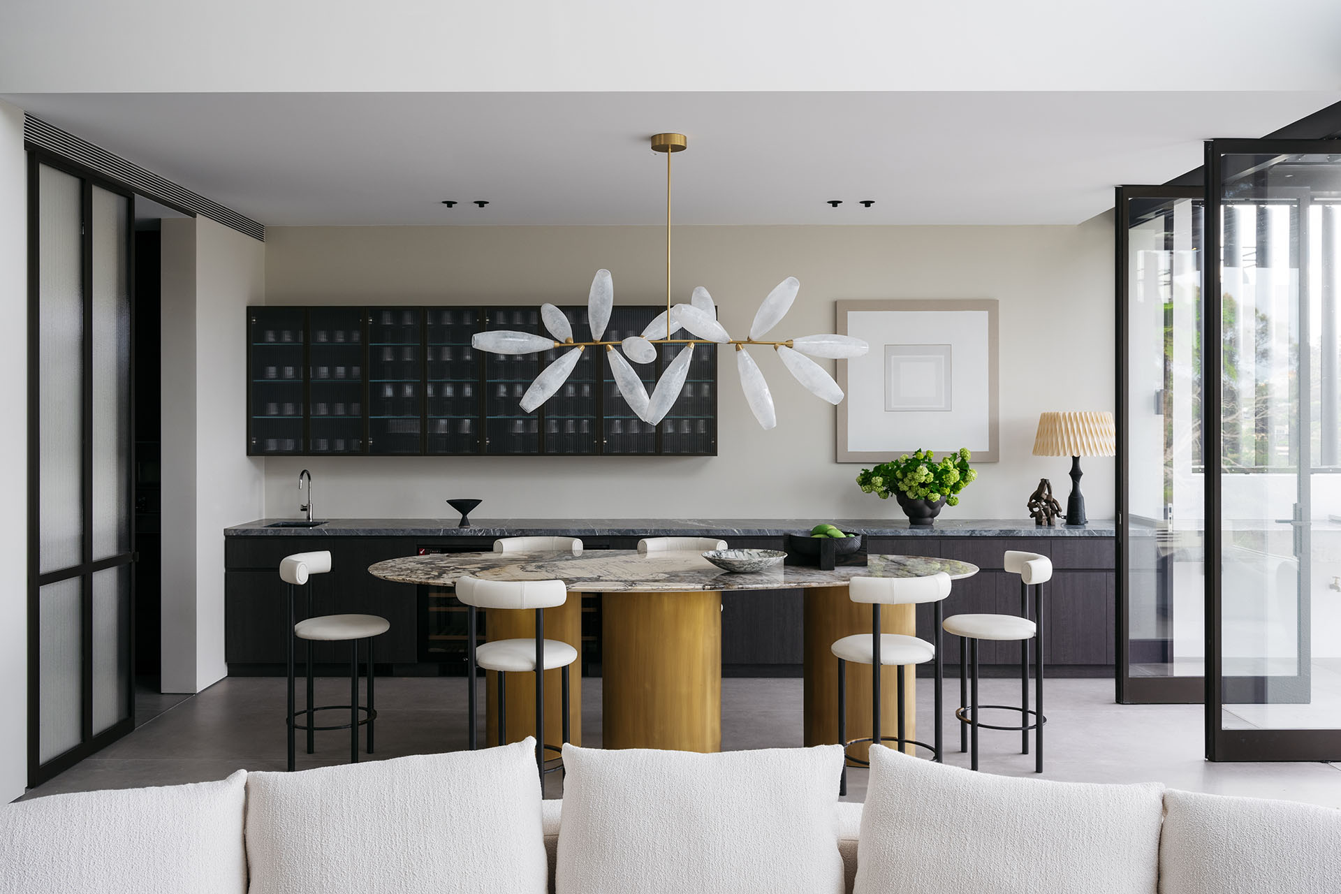 A modern kitchen designed by Nina Maya Interiors featuring a central island with a marble countertop, surrounded by custom white bar stools. A unique Gem Branch Chandelier by Giopato & Coombes hangs above.