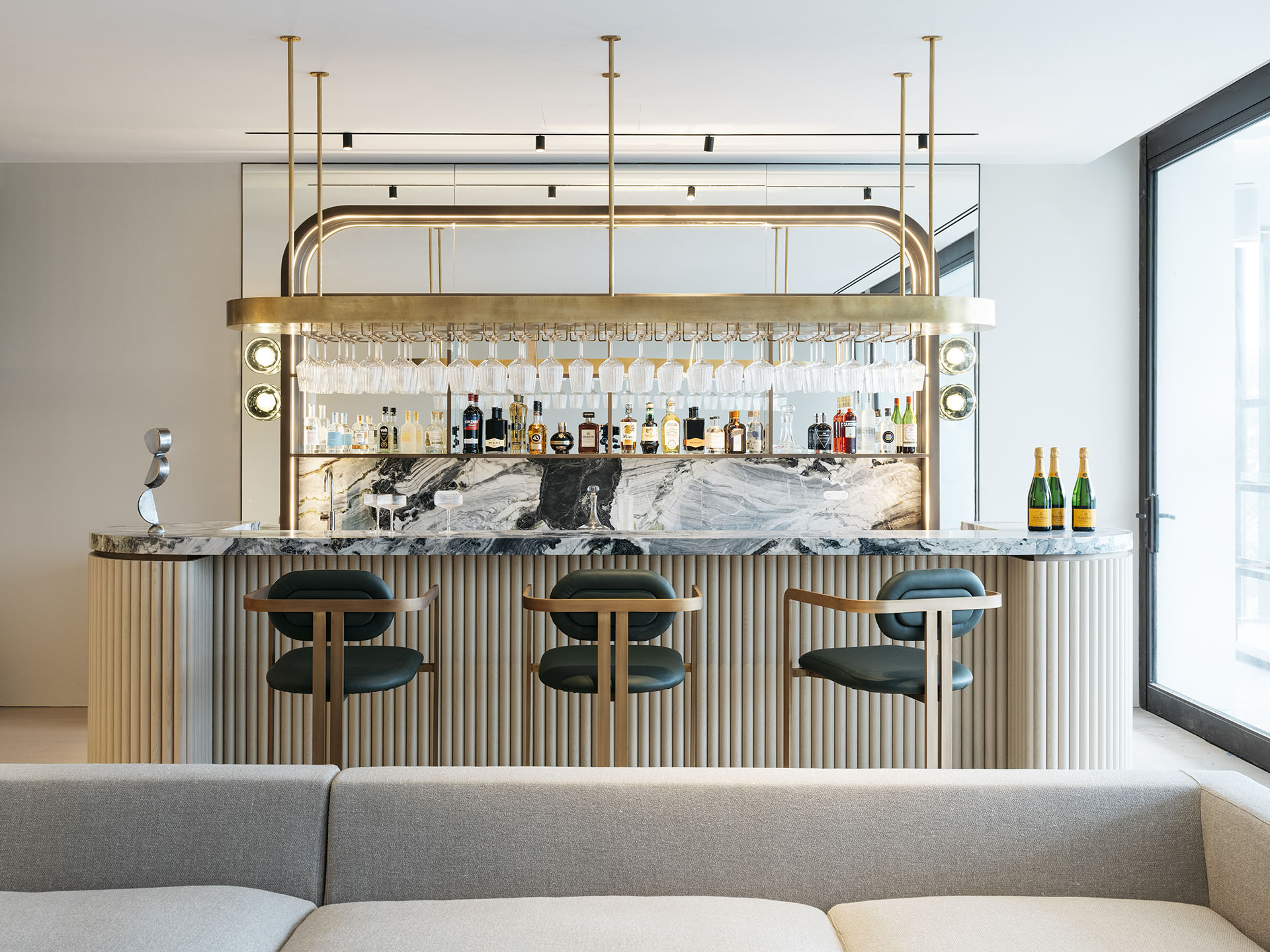 A luxurious home bar designed by Nina Maya Interiors with a marble counter, backlit shelves stocked with bottles, and green upholstered bar stools. This area showcases a blend of glamour and functionality.