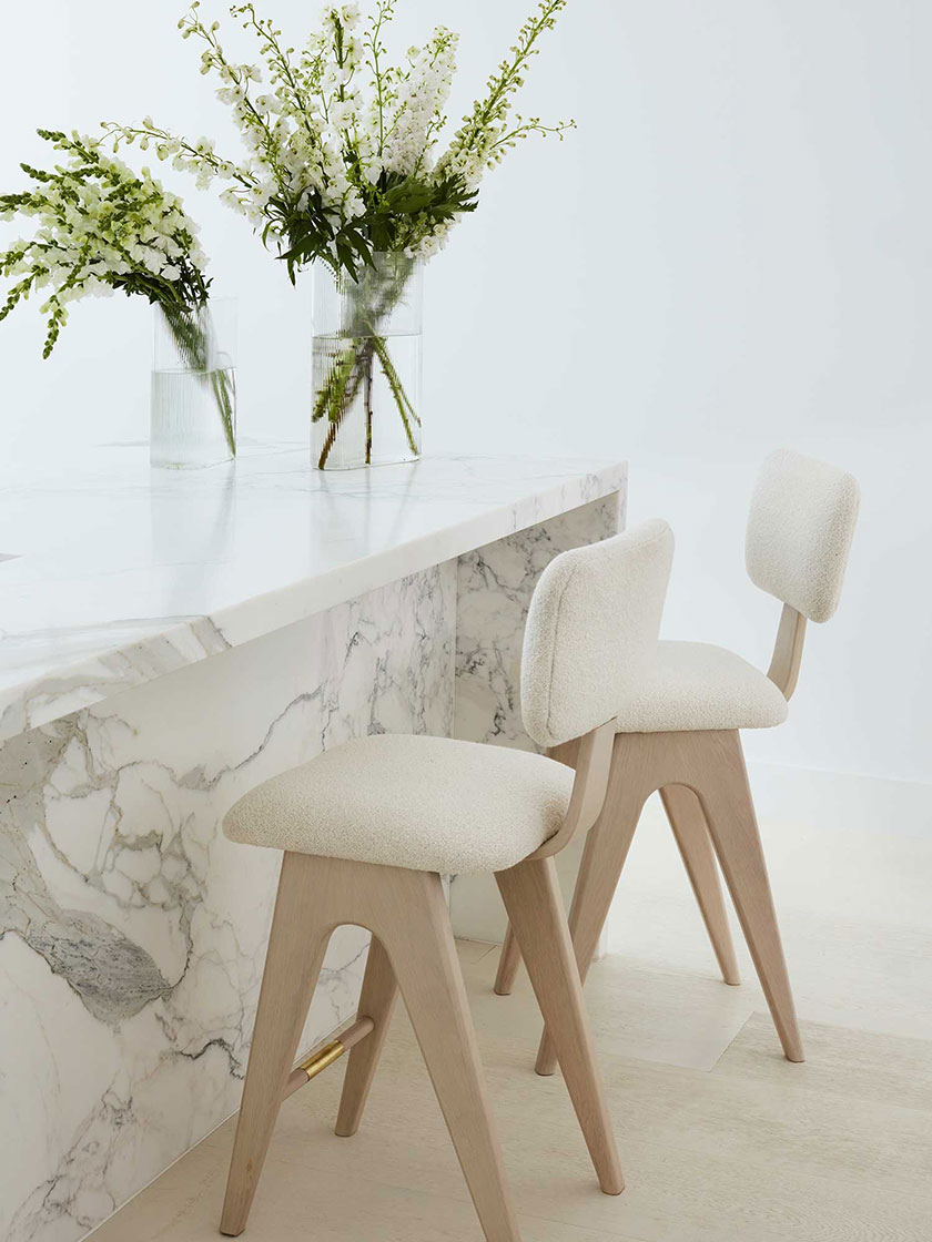 Elegant kitchen area by Nina Maya Interiors, with a marble counter, beige stools, and glass vases filled with white flowers, highlighting clean lines and minimalist design.