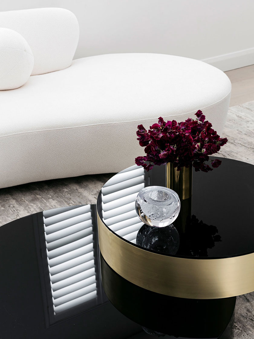 Close-up view of a gold and black coffee table with a glass ornament and flowers, white curved sofa in the background, designed by Nina Maya Interiors.